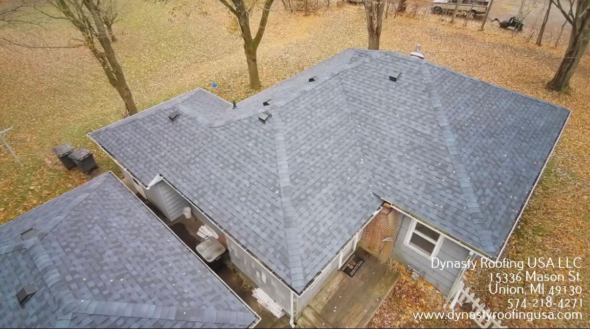 An elevated, aerial view of a gray shingled roof on a house, surrounded by a yard with scattered autumn leaves.
