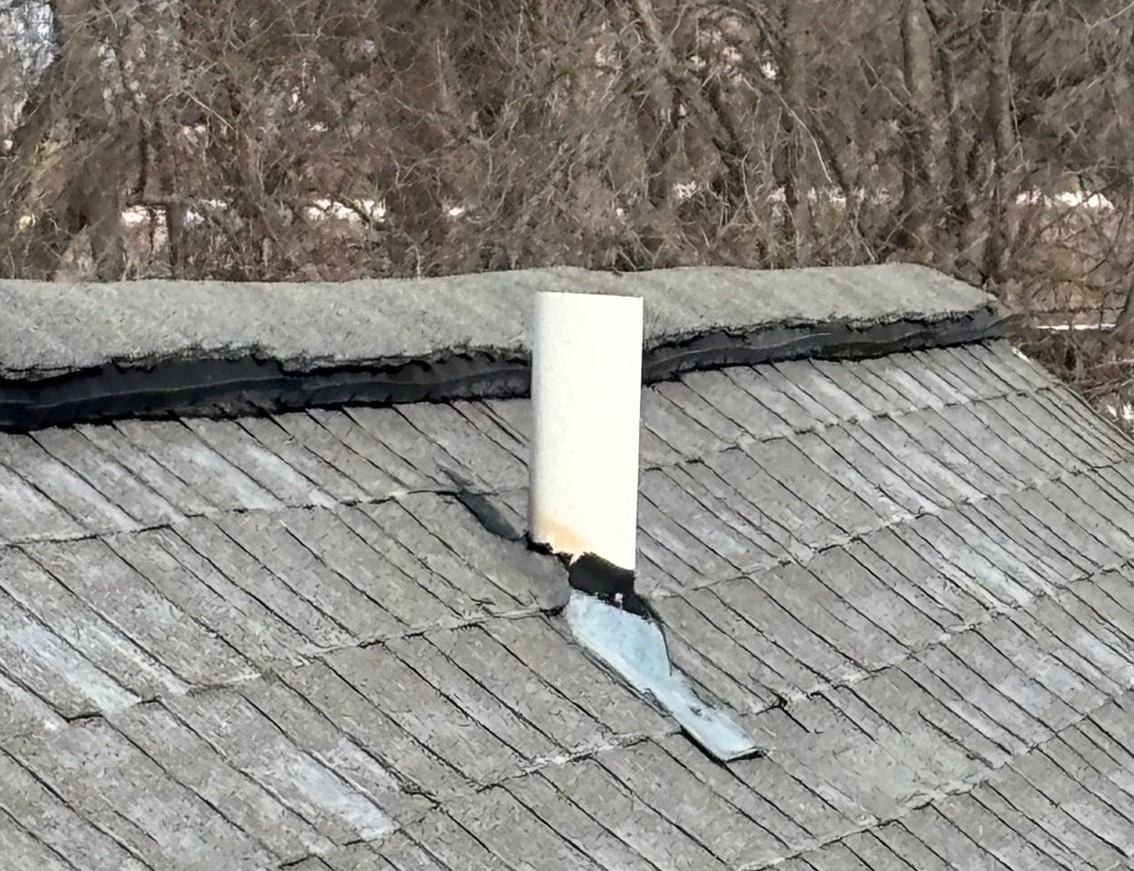 White chimney pipe on a gray shingled roof with damaged flashing and a loose metal panel.