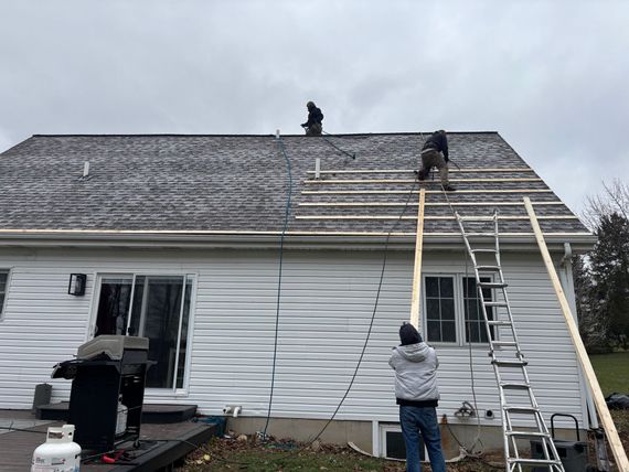 Shingle to metal roofing replacement Workers replacing a shingle roof with a metal roof on a white house roof, with ladders and roofers visible.