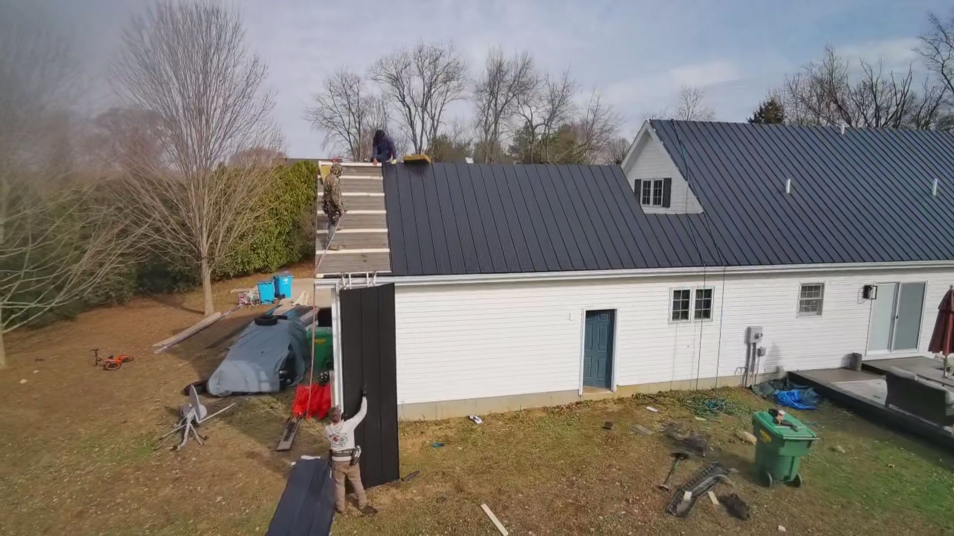 Two workers on a house roof with extension ladders leaning against the siding and a patio below