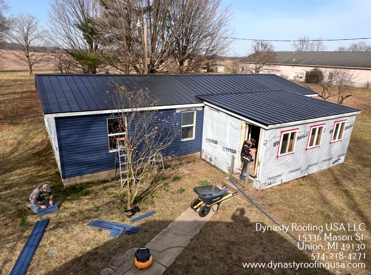 Workers installing a dark metal roof on a small blue house, with building materials scattered on the lawn.