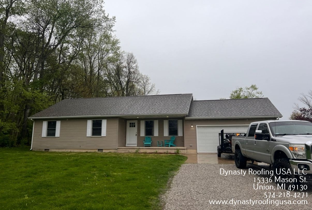 A single-story tan home with gray shingles and a white garage, featuring a pickup truck parked on a gravel driveway.