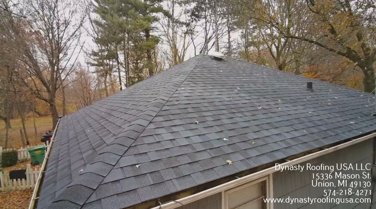 A high-angle view of a grey shingled residential roof with trees in the background and branding text in the corner.