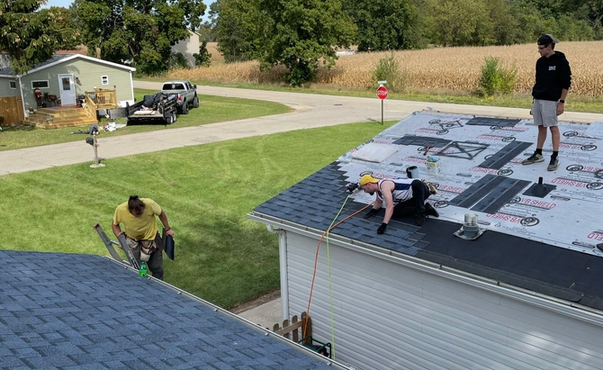 Roofer installing dark gray shingles on a roof using a nail gun on a sunny day.