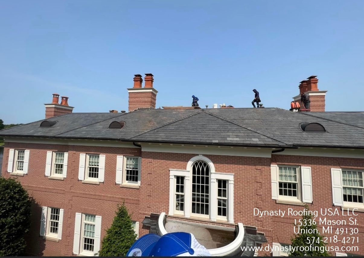 Workers on a red brick house roof with chimneys under a blue sky.