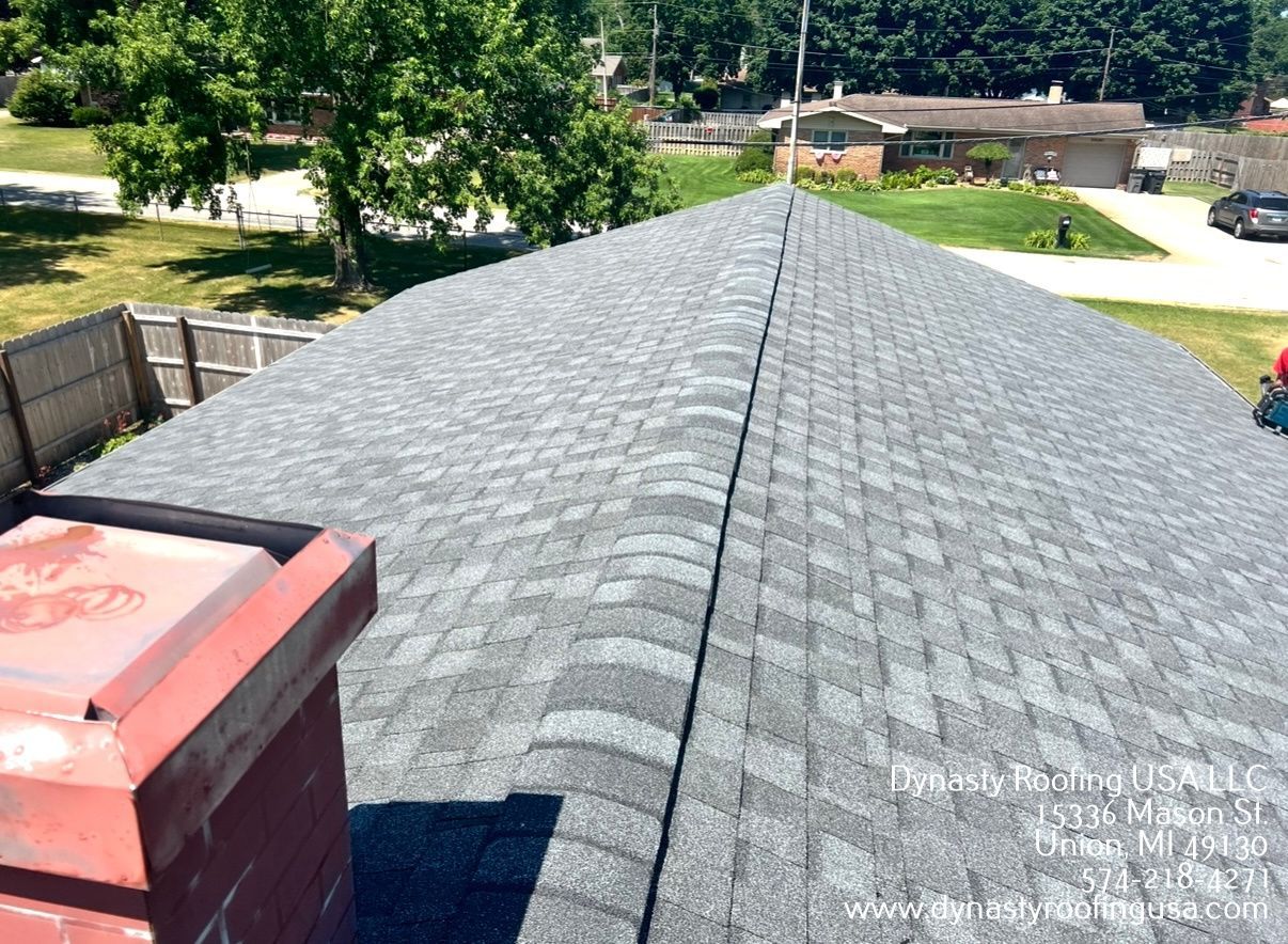 Gray shingled roof with a red chimney, trees, and houses in the background on a sunny day.