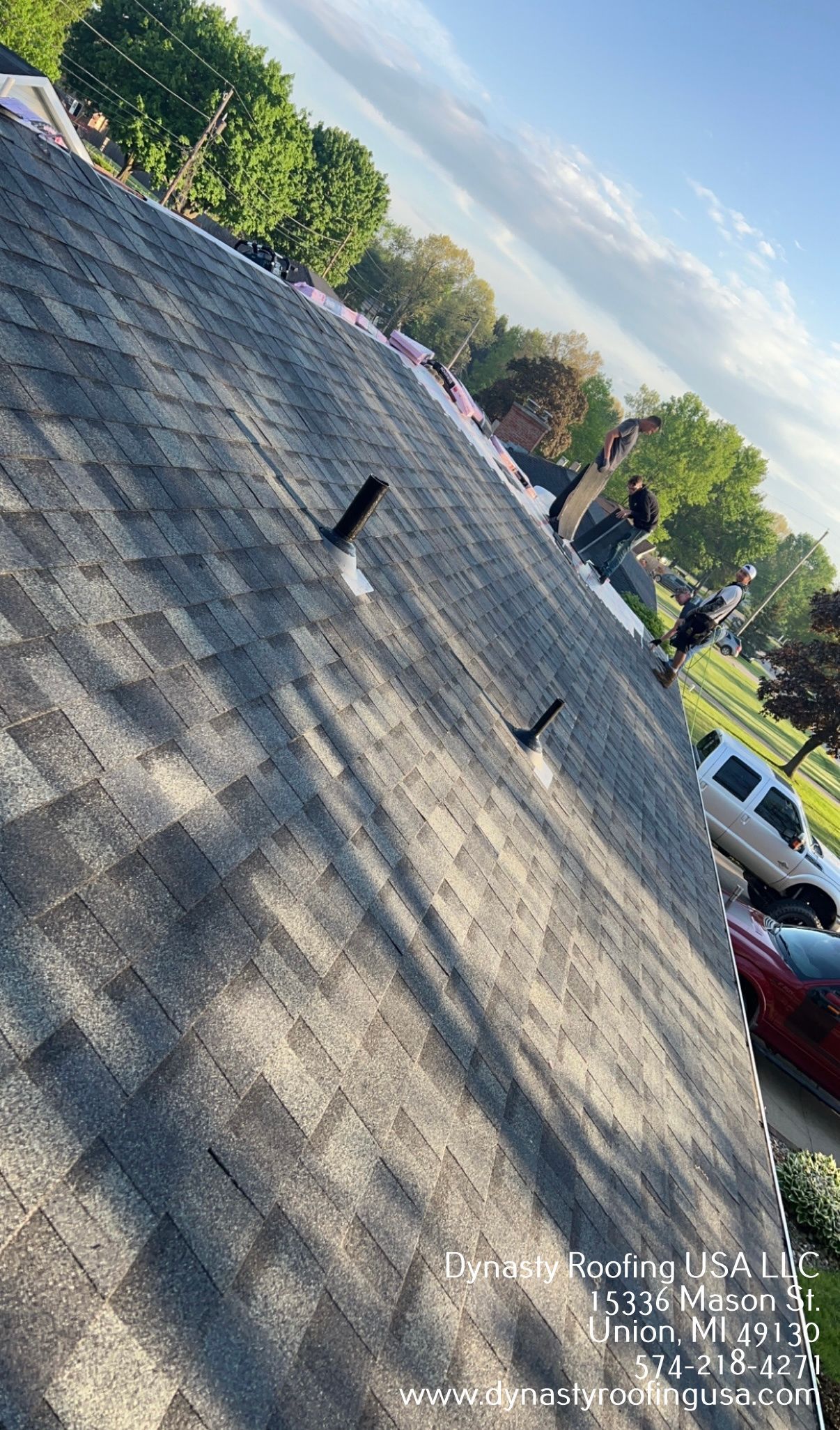 A shingled roof with a shadow of someone standing on it, trees and cars are in the background.