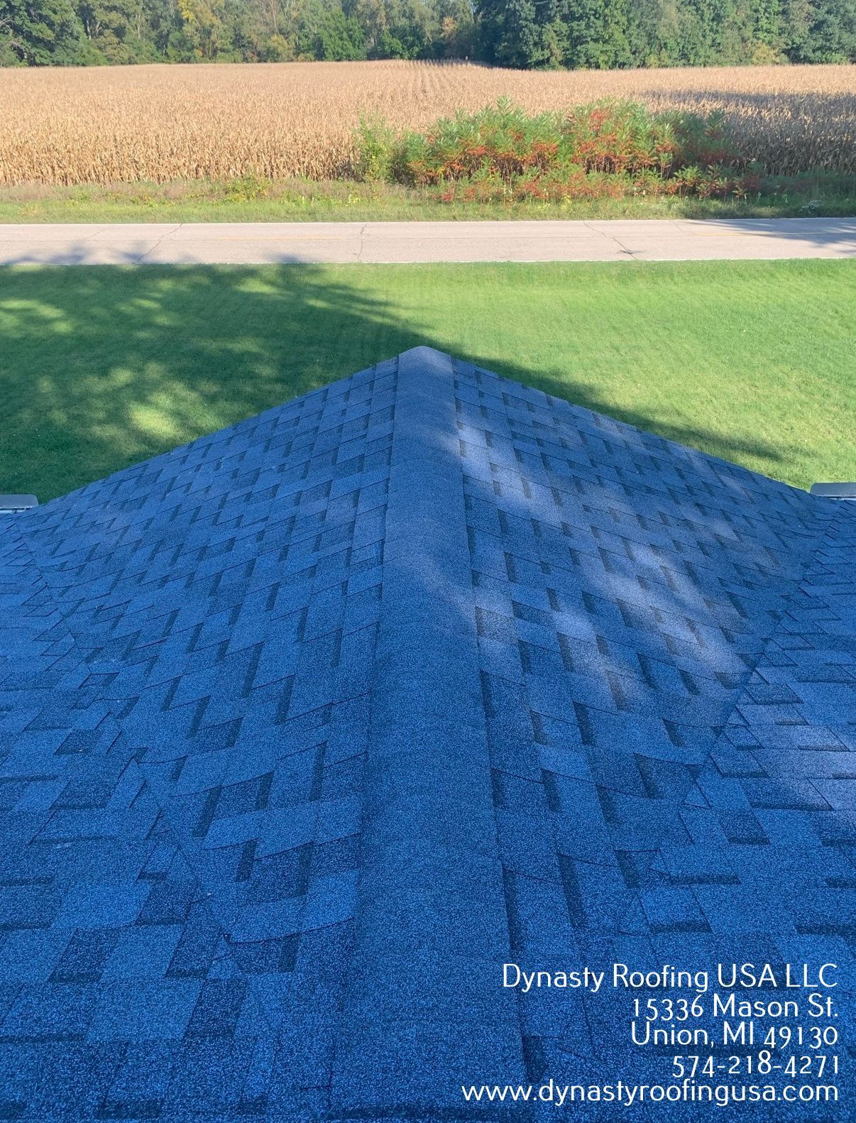 Blue shingled roof with a green lawn and field in the background. Dynasty Roofing USA LLC logo.