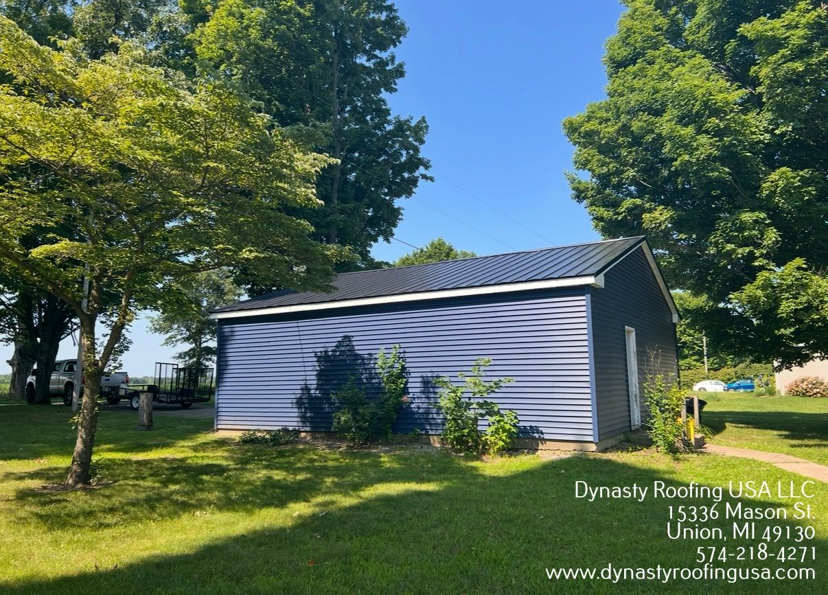 Blue-sided building with dark roof in a grassy yard, trees in the background, blue sky.
