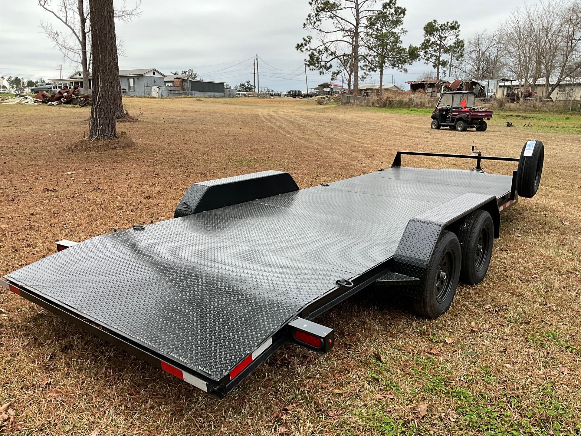 A black diamond-plated steel car hauler trailer parked on a grassy field with a utility vehicle in the background.