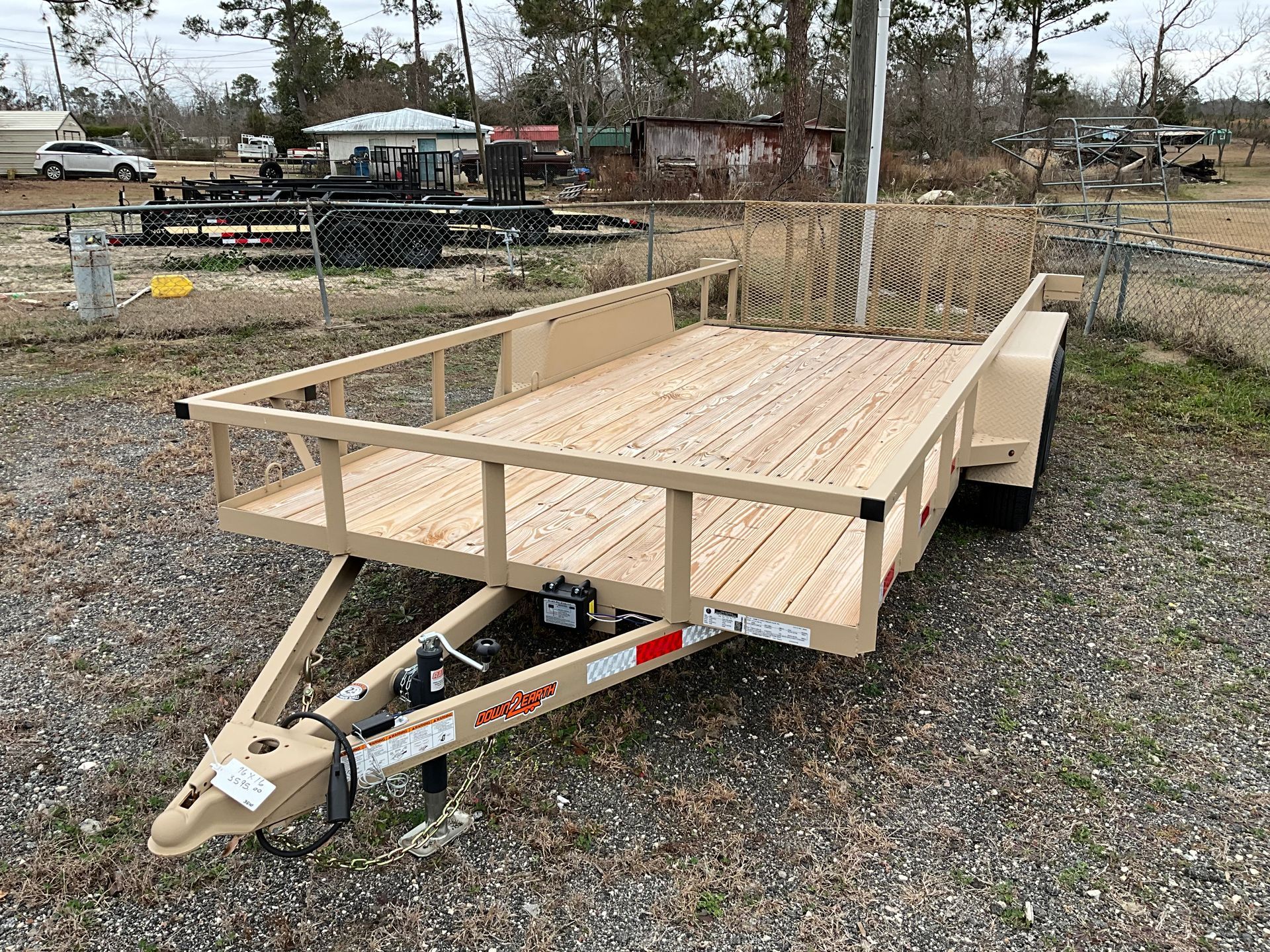 A new tan utility trailer with wooden floorboards and a rear mesh gate parked on a gravel lot.