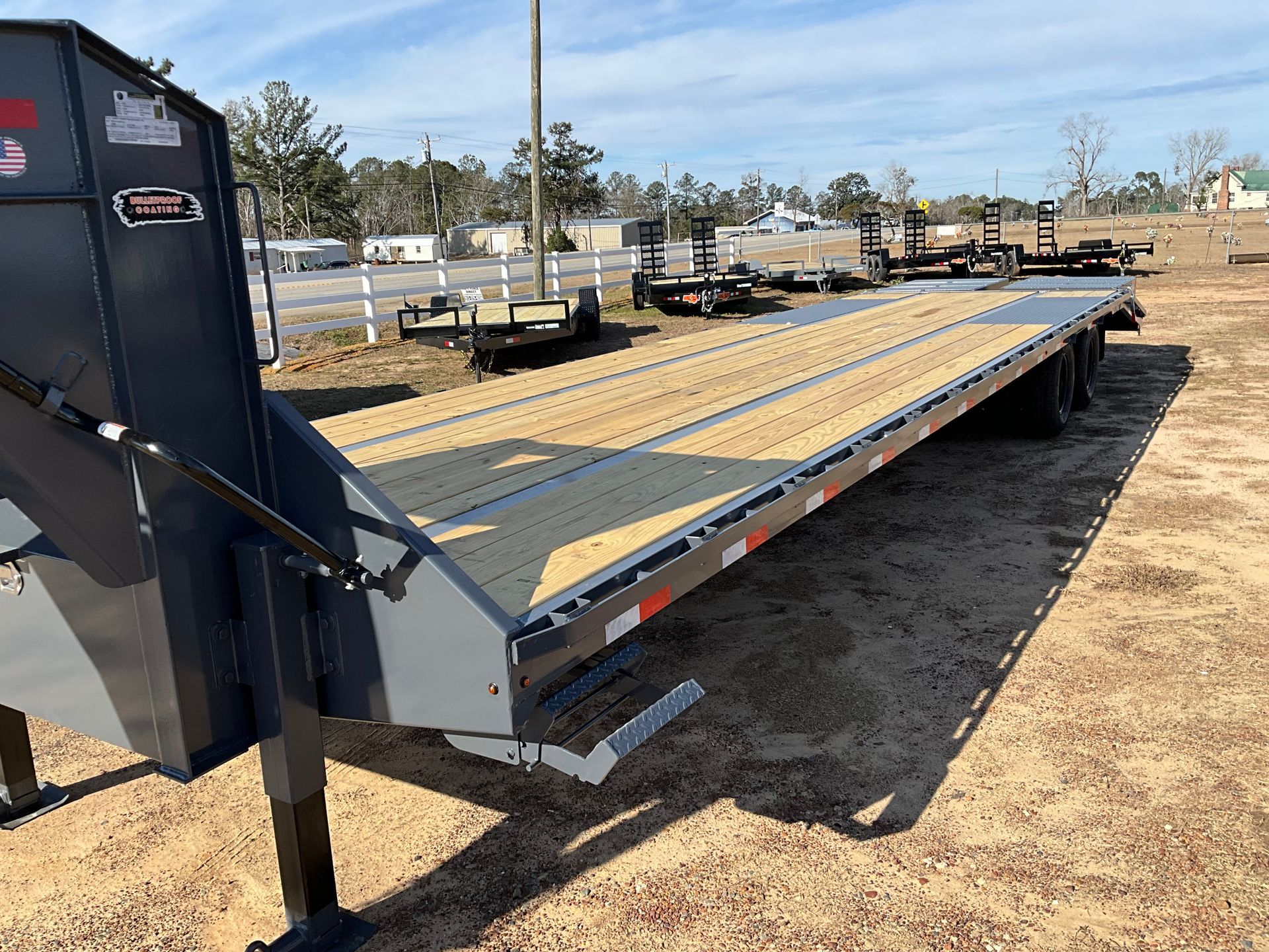 A dark gray flatbed trailer with a wooden deck parked on a dirt lot on a sunny day.