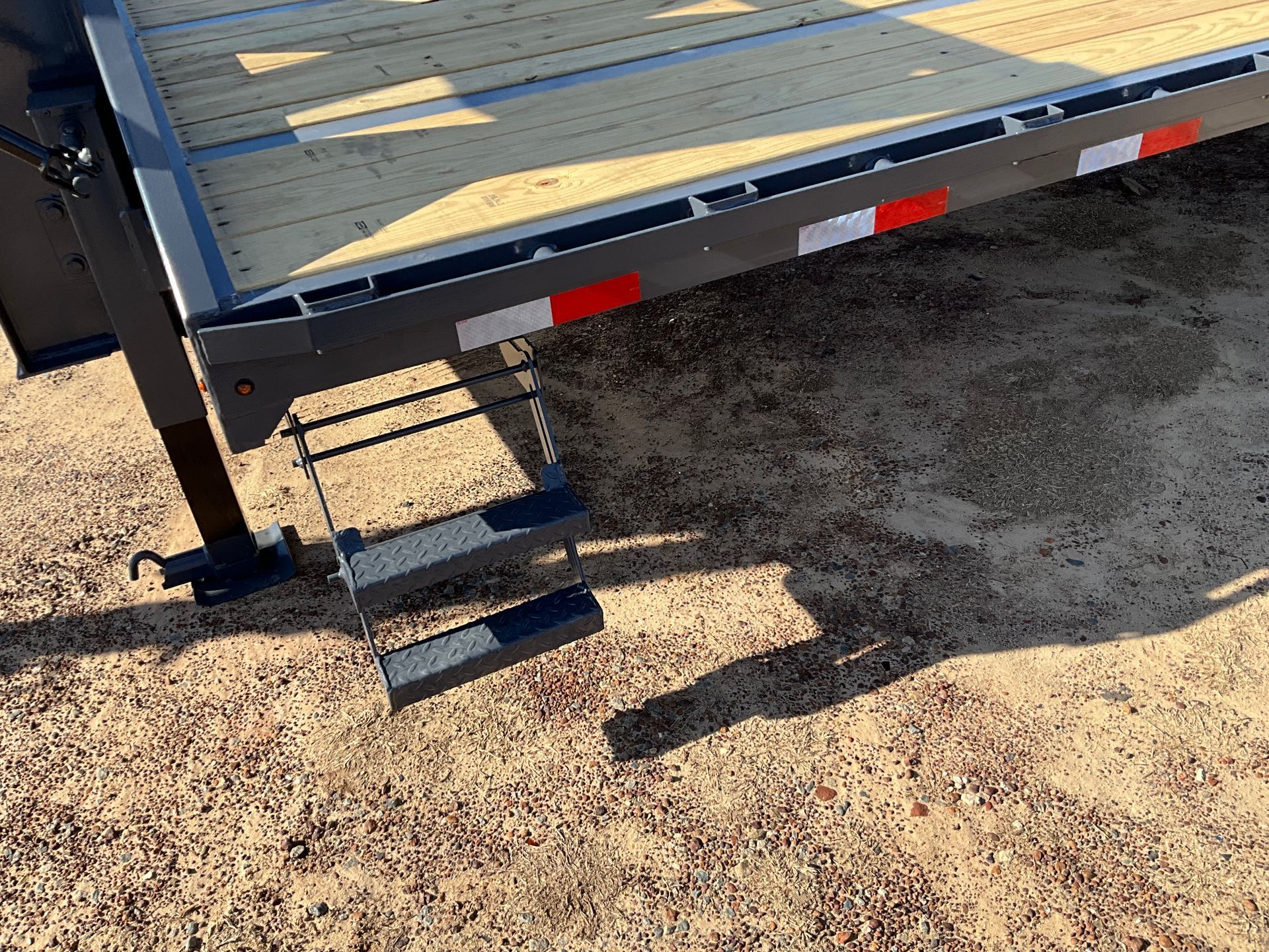 Fold-down metal steps attached to the side of a trailer with a wooden deck, parked on a dirt lot.