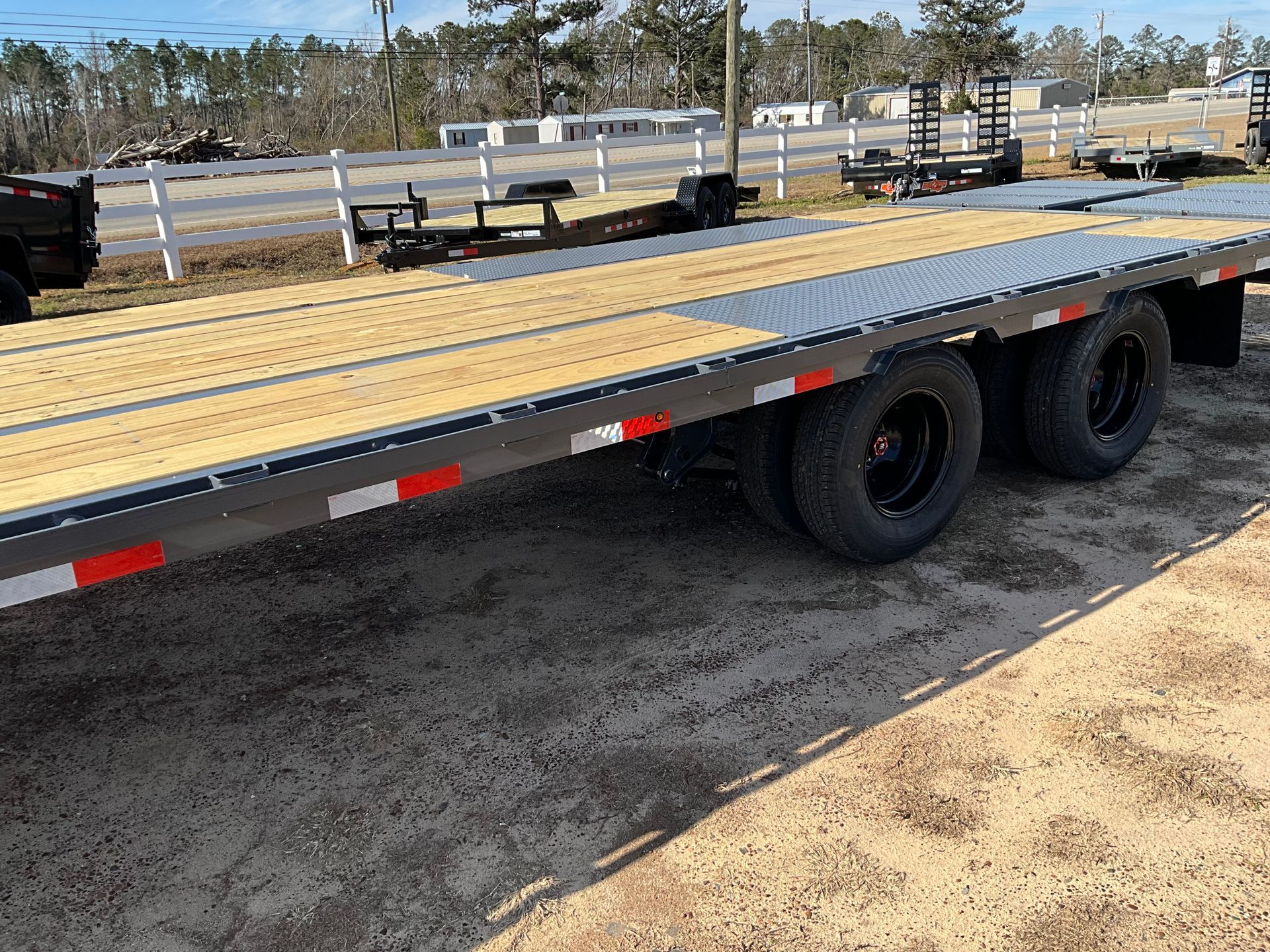 A flatbed trailer with a wooden deck and black wheels parked on a gravel lot in front of a white fence.