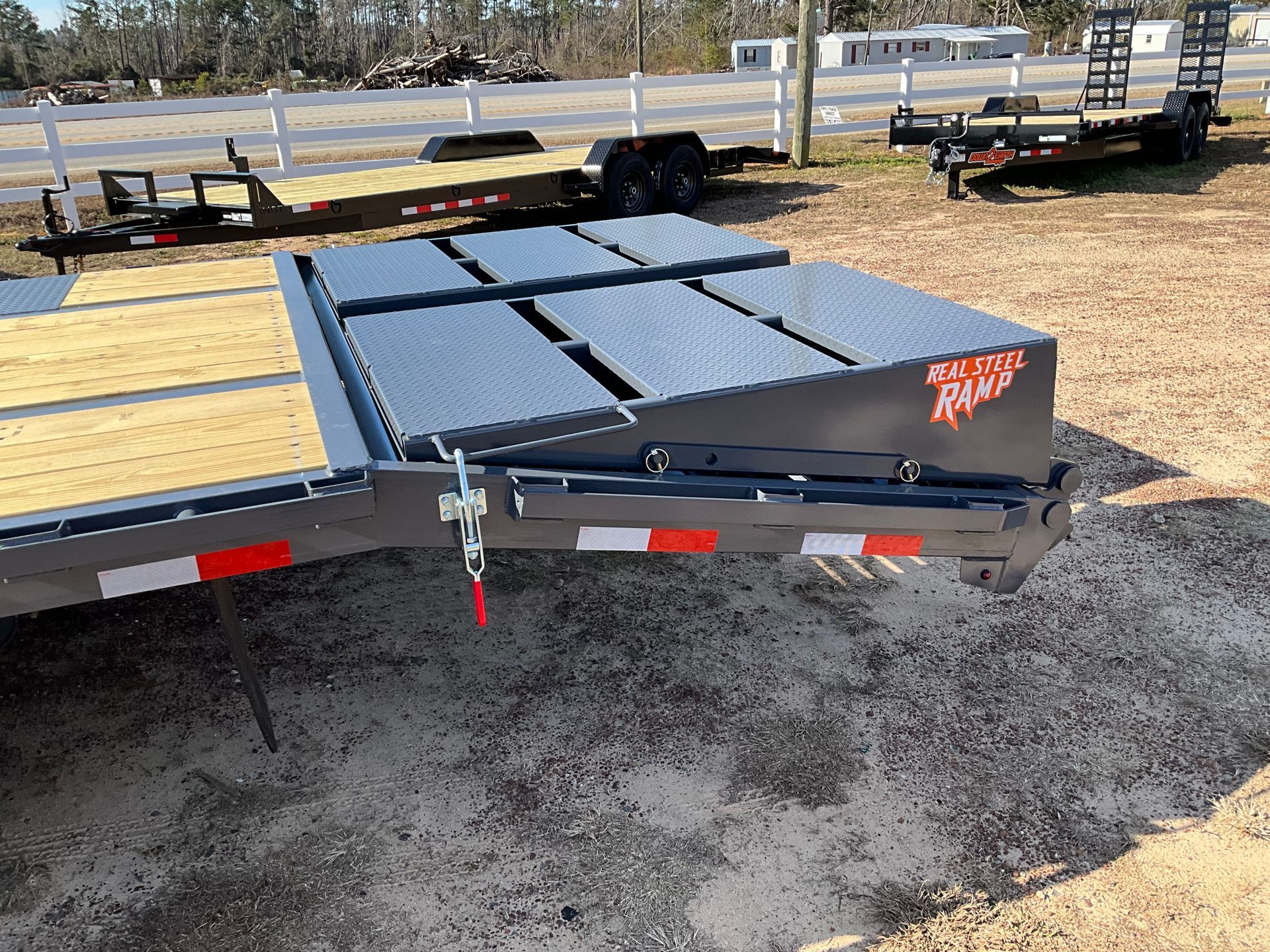 A close-up of a trailer's metal diamond-plate ramp system, folded flat on the wooden deck in an outdoor lot.