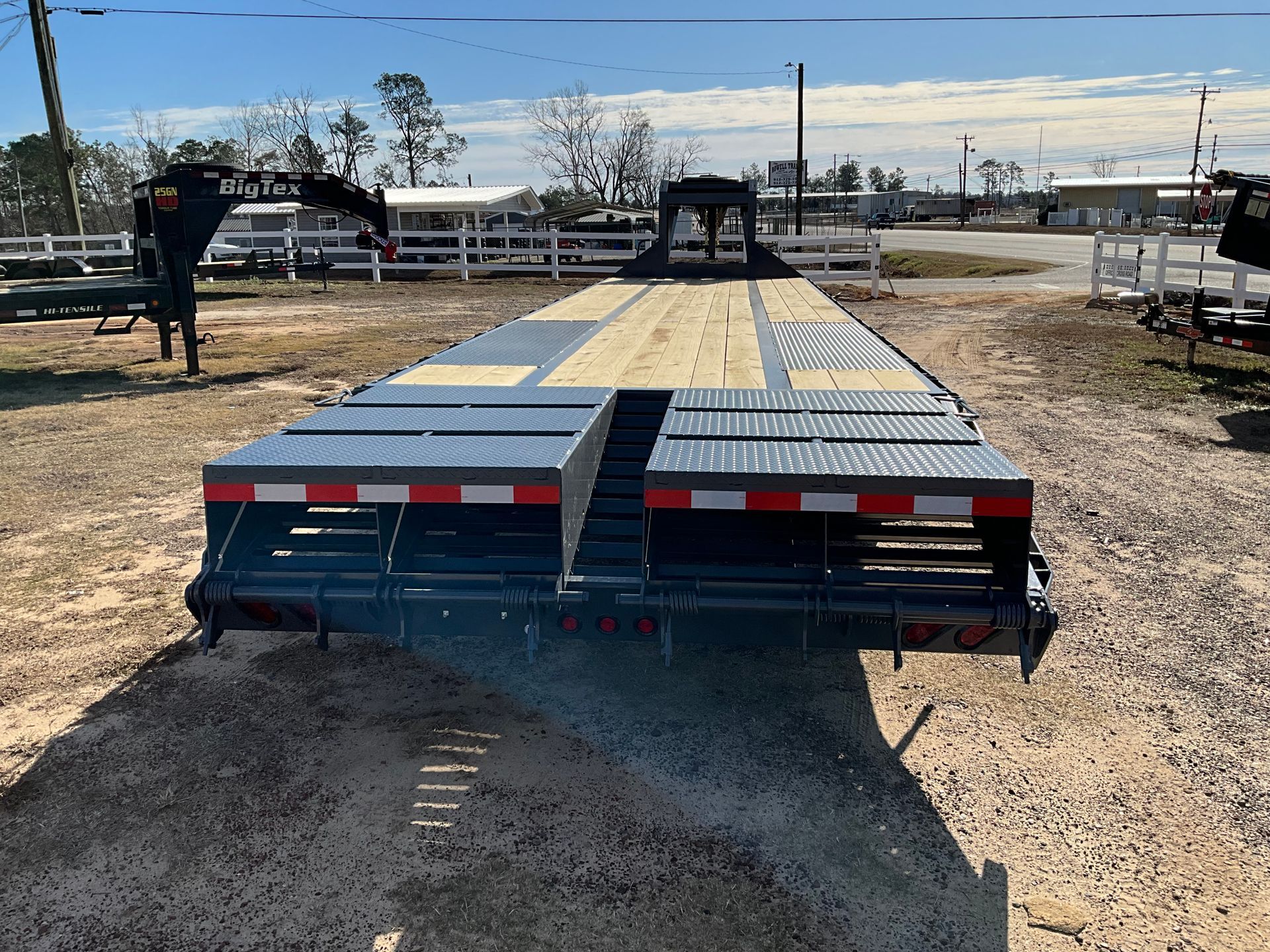 A dark gray Big Tex flatbed trailer with a wooden deck and metal ramps, parked on a dirt lot on a sunny day.
