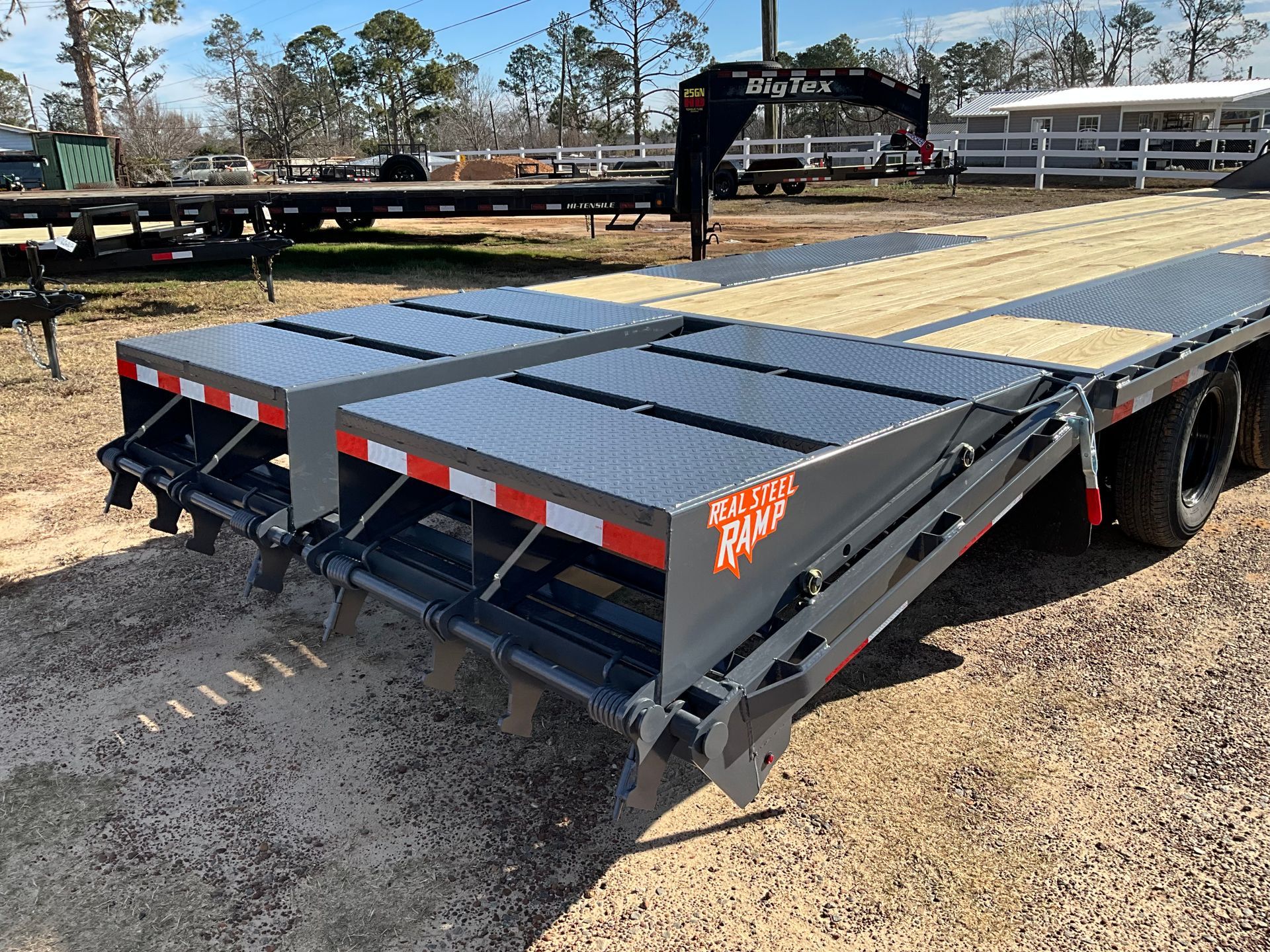 Rear view of a grey heavy-duty trailer with diamond-plate ramps and wooden decking parked on a gravel lot.