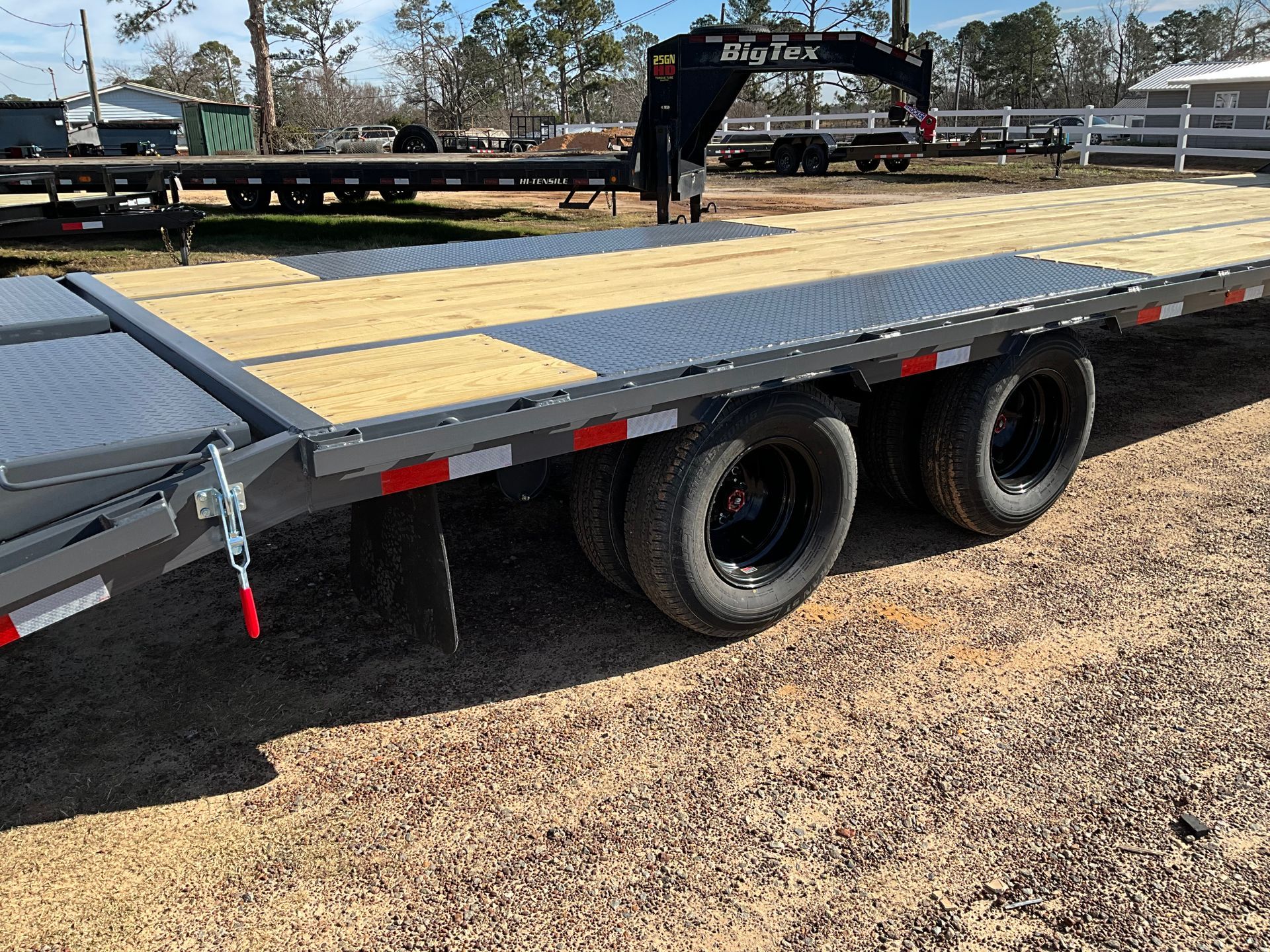 A gray flatbed gooseneck trailer with a wooden deck and dual wheels parked on a gravel lot under a sunny sky.