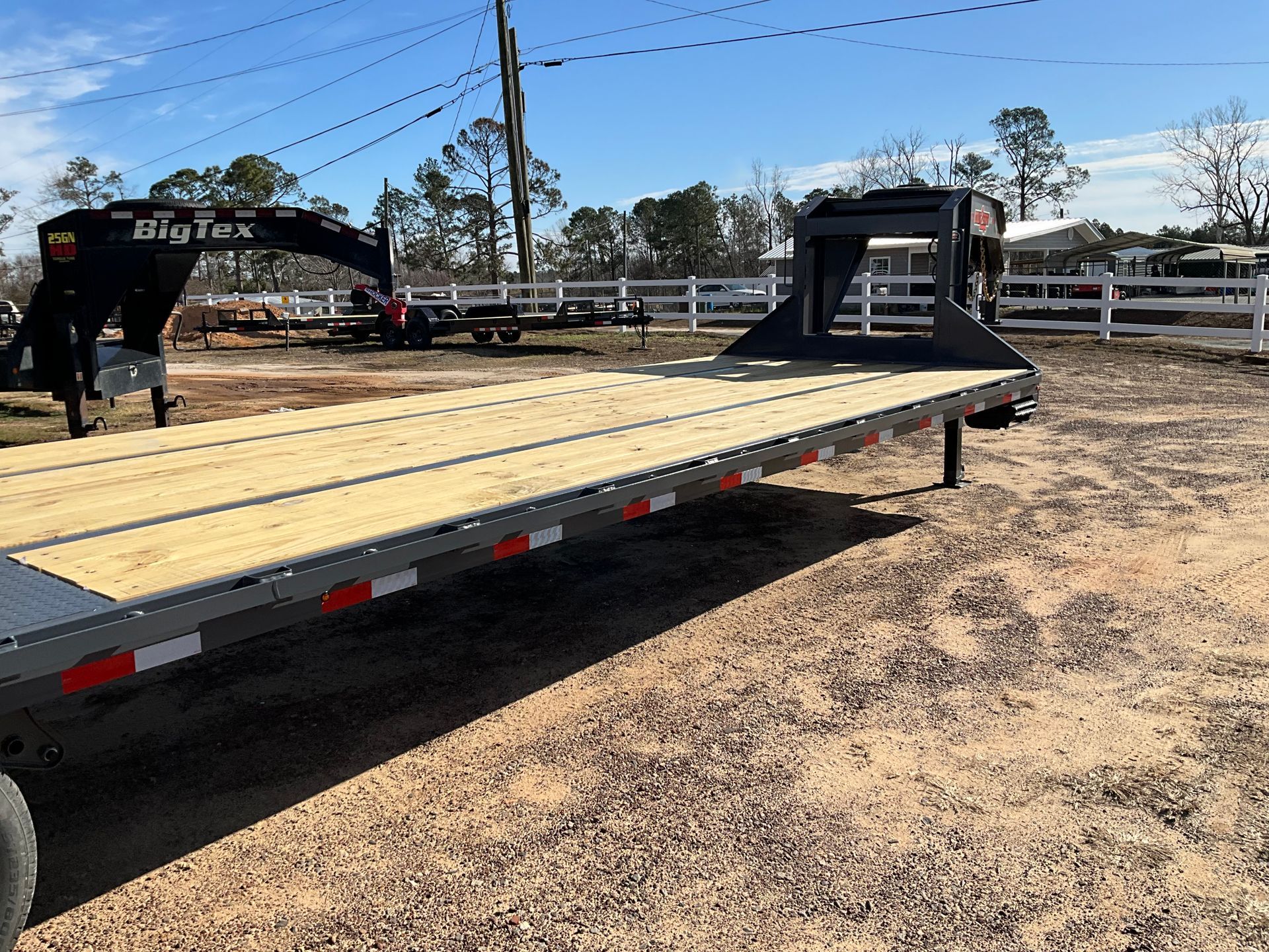 A flatbed gooseneck trailer with a wooden deck and a black metal frame parked on a dirt lot on a sunny day.