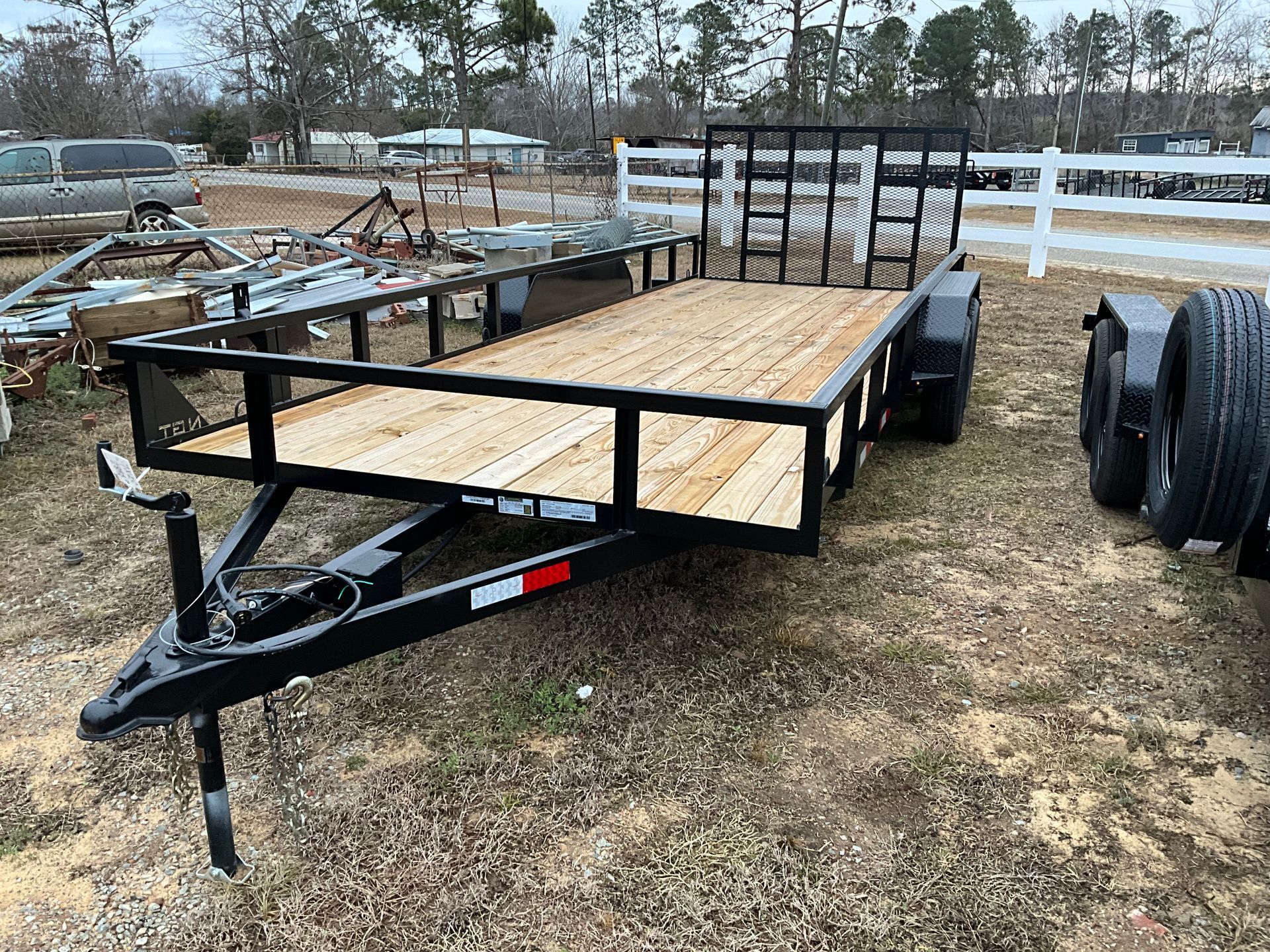 A black utility trailer with a new wooden deck and a rear gate, parked on a gravel lot next to a white fence.