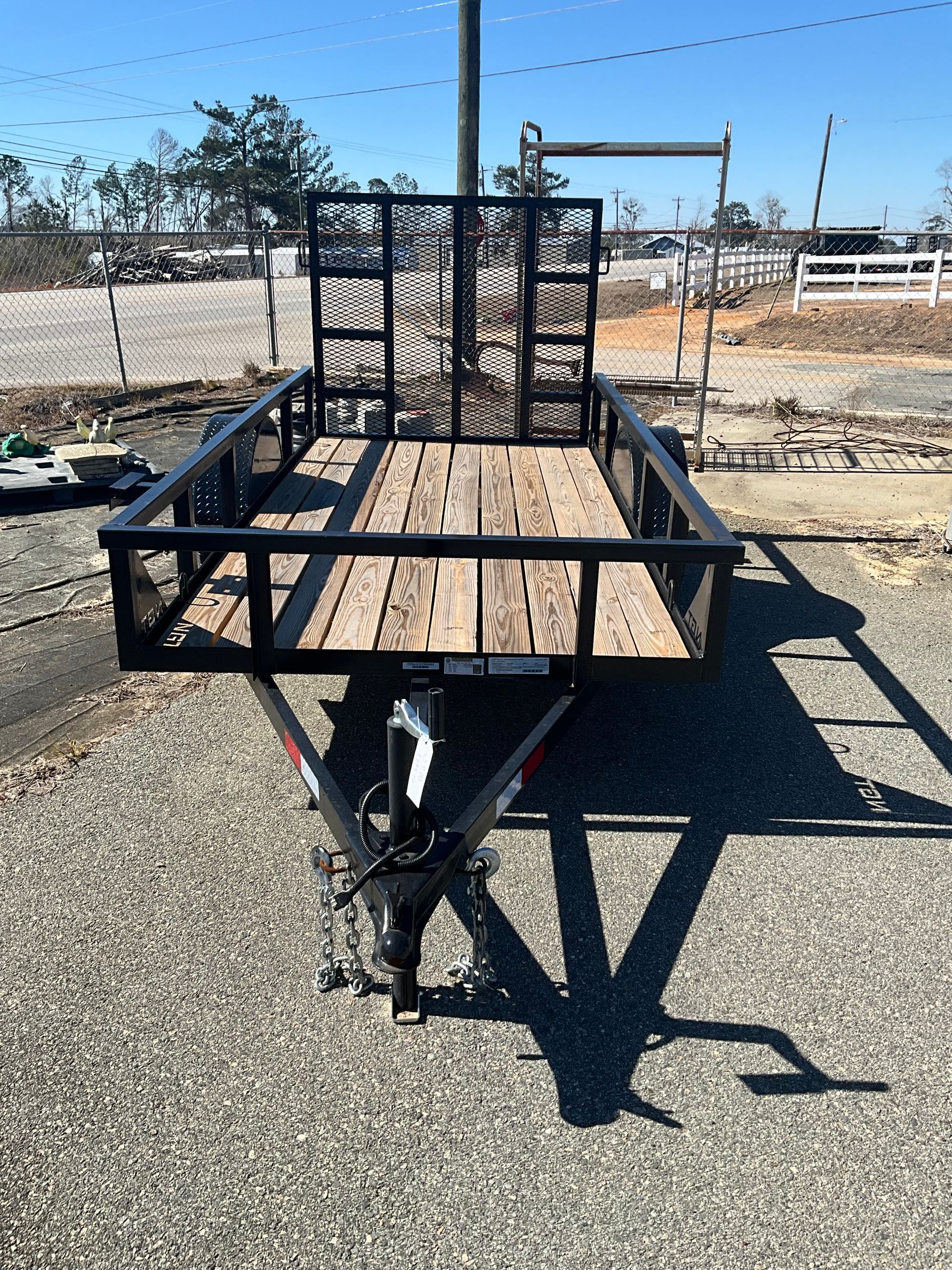 A black utility trailer with a wooden floor and rear mesh gate parked on a gravel surface outdoors.