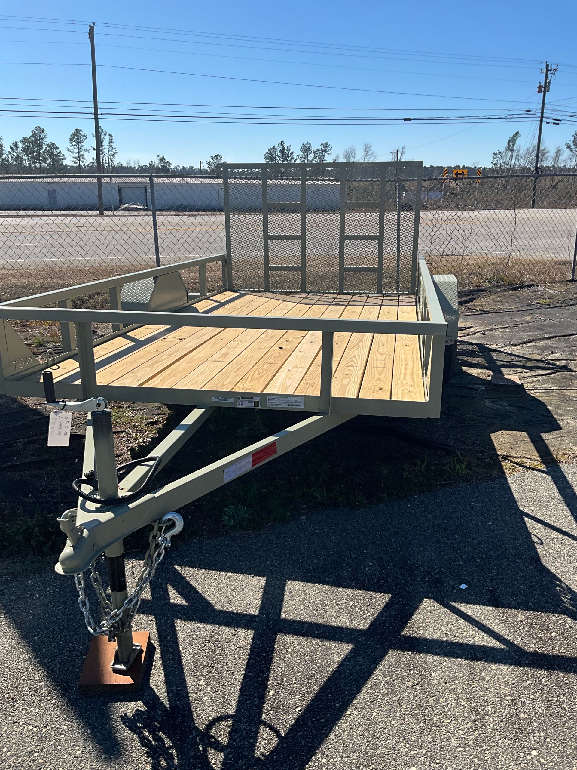 A green utility trailer with a wooden floor sits on a gravel lot under a bright blue sky.