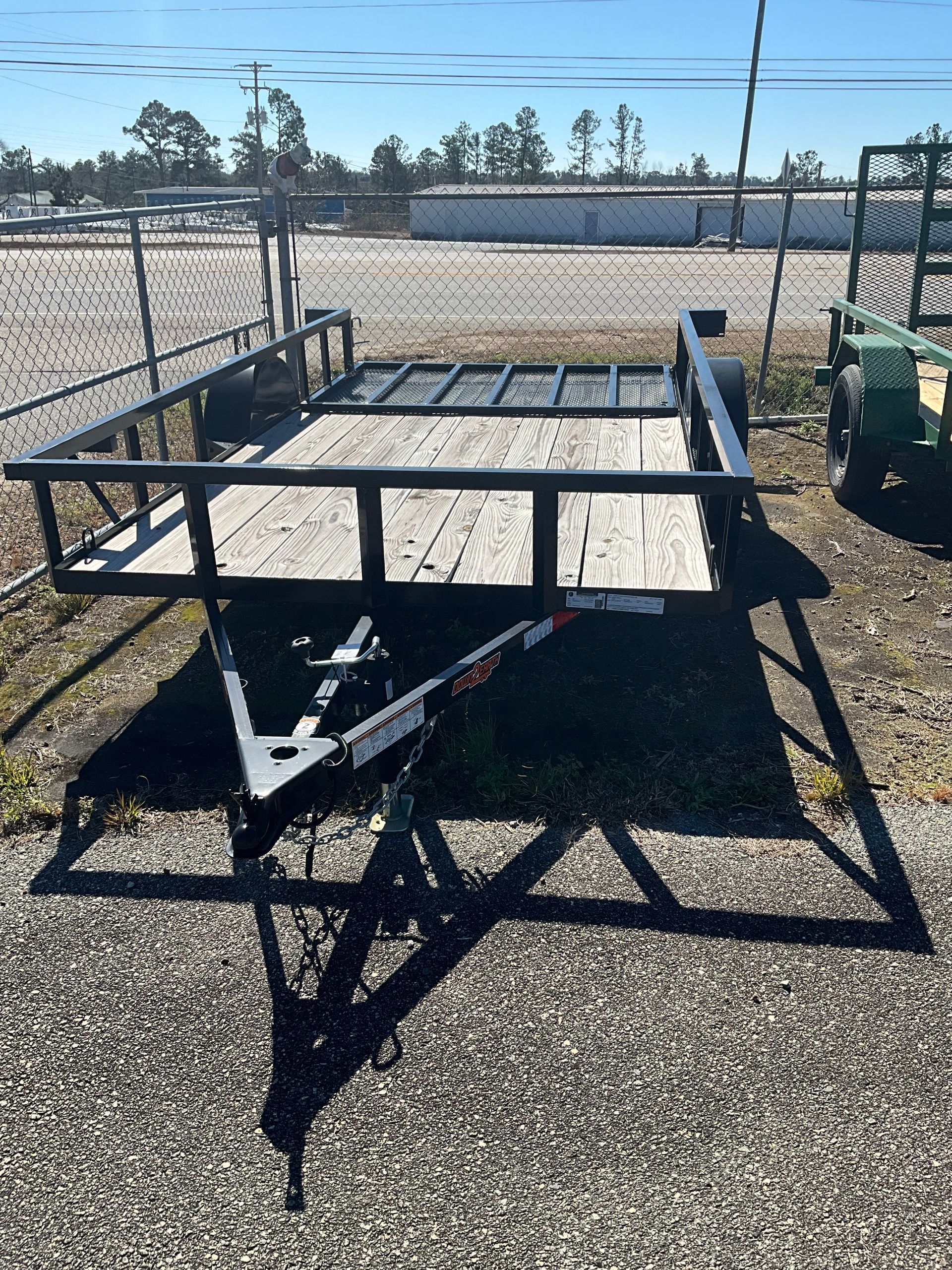 An empty black metal utility trailer with a wooden floor sits on a gravel lot under a clear blue sky.
