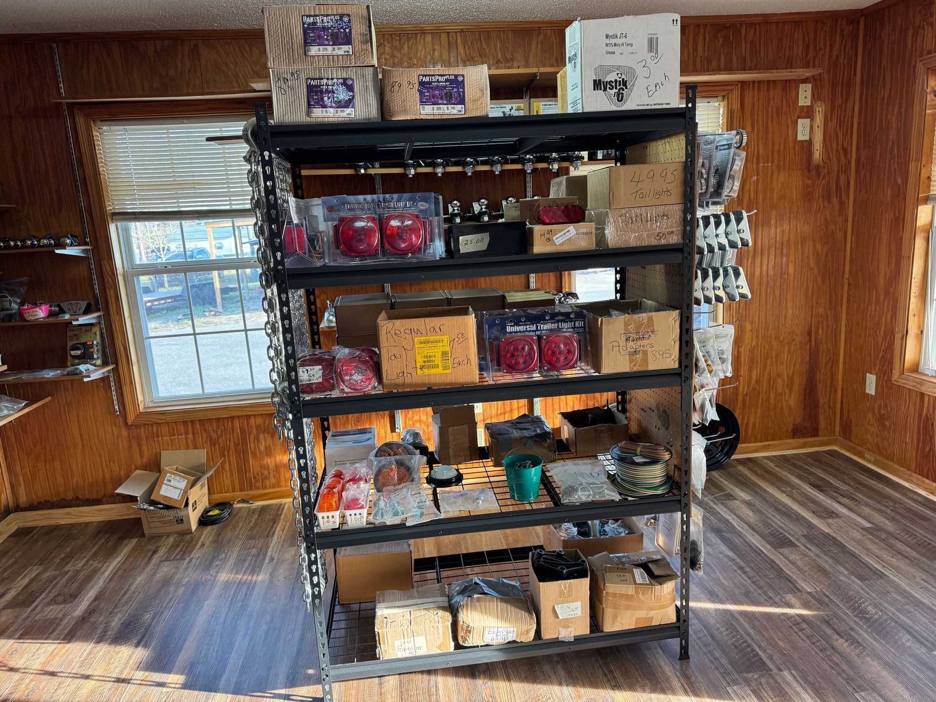 A dark metal shelving unit in a wood-paneled room is filled with various boxed and bagged automotive or hardware parts.