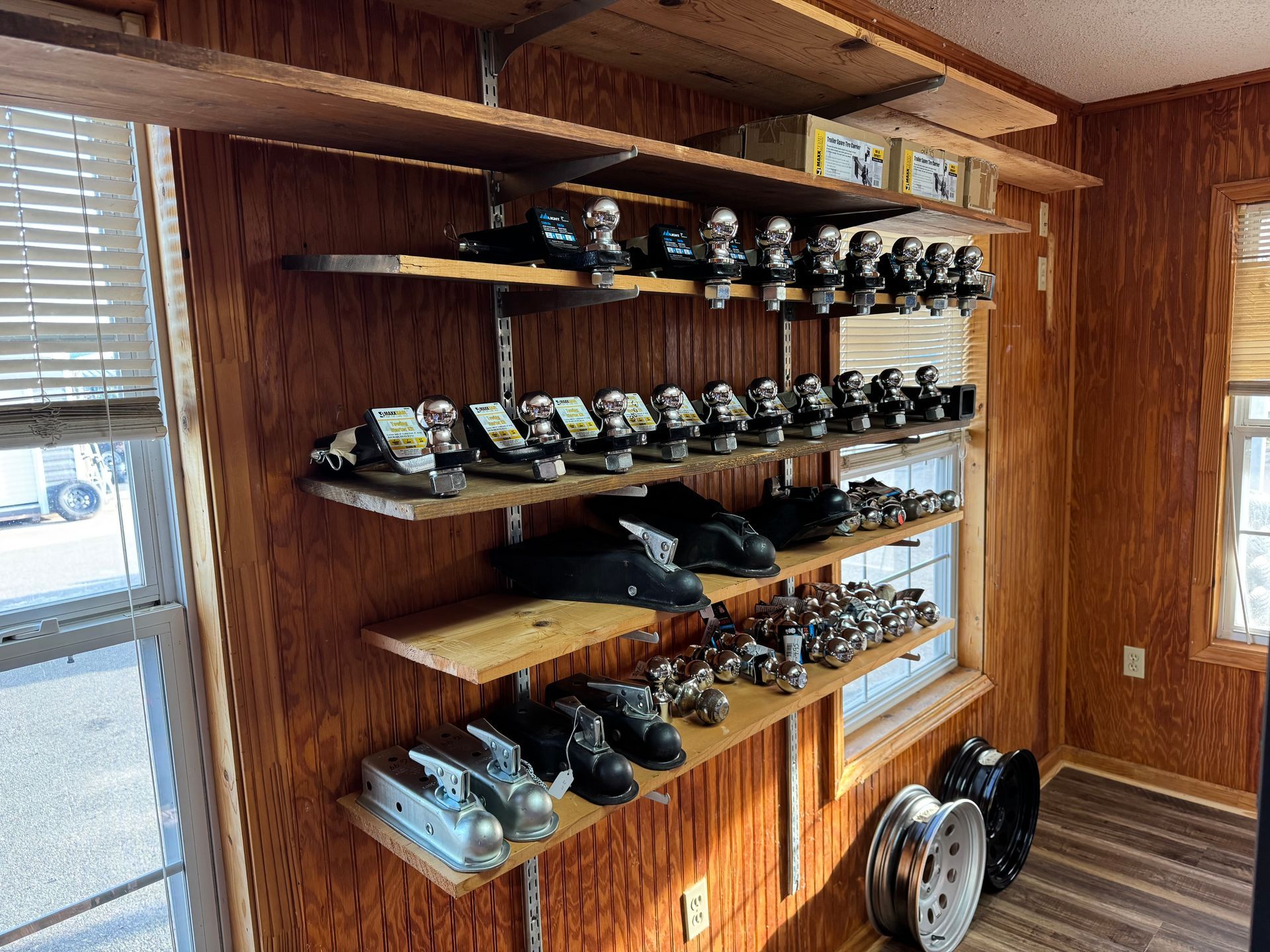Shelves in a room with wood-paneled walls display an assortment of trailer hitches and two spare wheels on the floor.