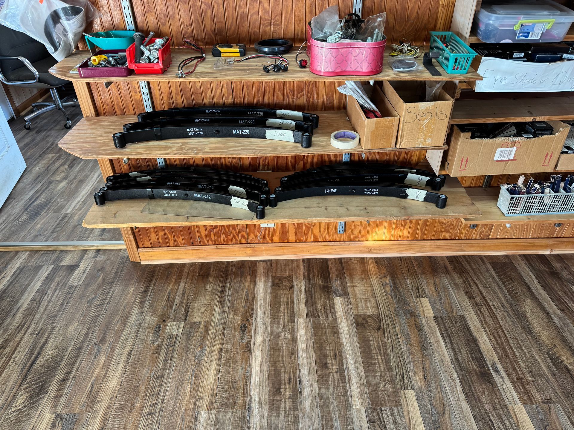 Black leaf springs stored on wooden shelves in a workshop with bins of parts and a desk chair.