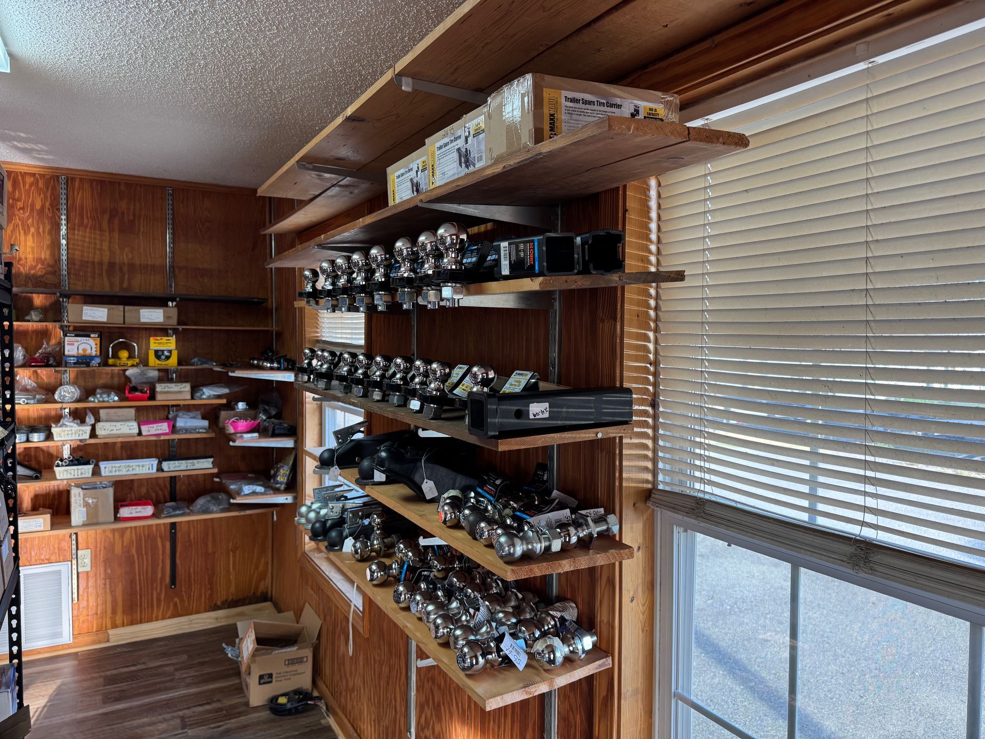 Wooden shelving units inside a room, stocked with numerous metallic trailer hitch balls, beside a window with blinds.