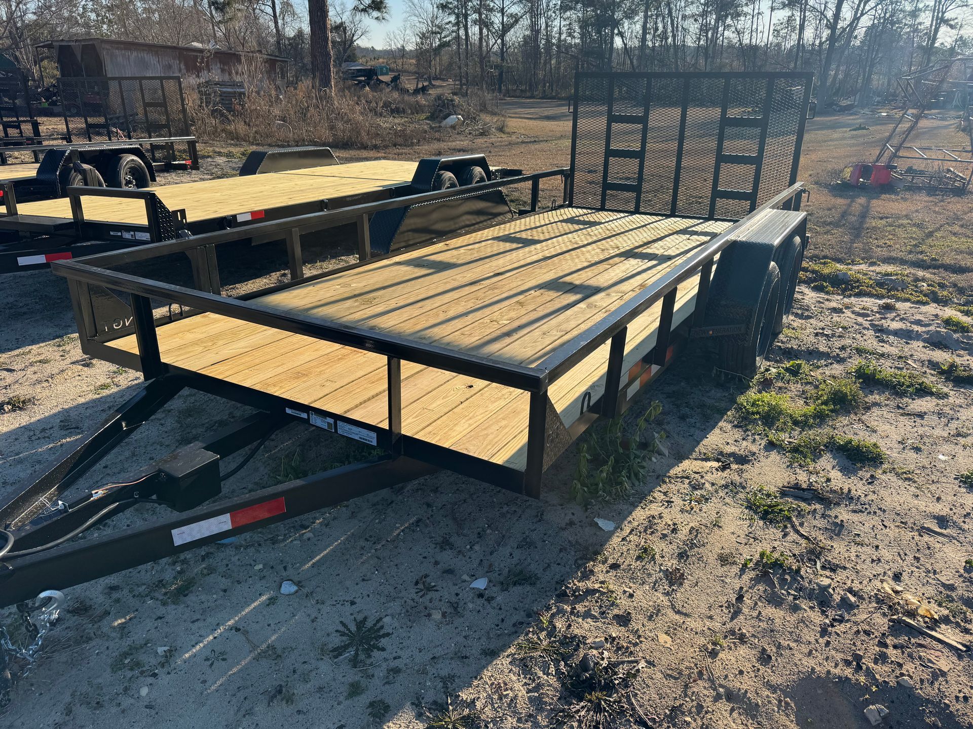 A black metal utility trailer with a new wooden deck and a rear ramp, parked on a dirt lot on a sunny day.