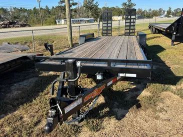 A black flatbed equipment trailer with a wood deck and rear mesh ramps parked on a grass lot.