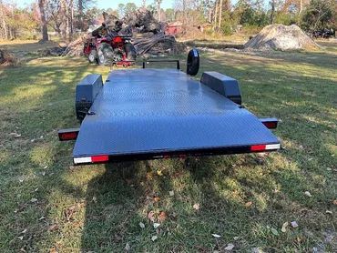 A black metal utility trailer sits on a grassy field with a large pile of debris in the background.