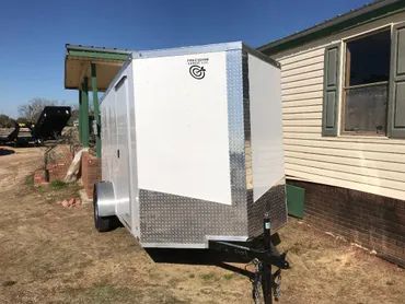A white enclosed trailer with a diamond-plate metal lower front, parked on grass beside a light-colored mobile home.