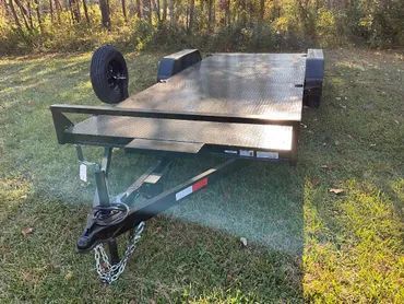 A flatbed utility trailer with diamond-plate steel decking, parked on a grass lawn near a wooded area.