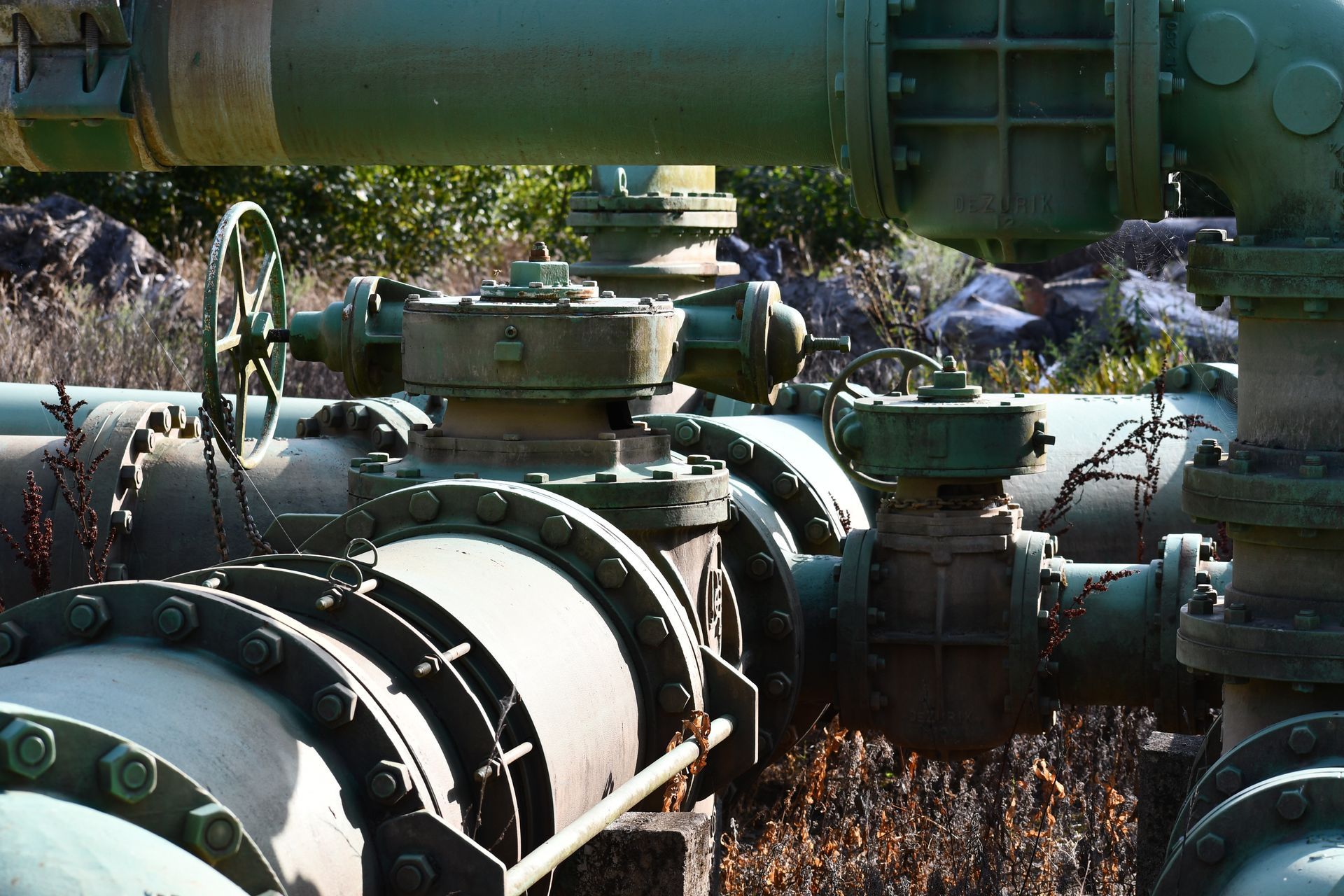 Industrial green pipelines and valves in an outdoor utility system infrastructure for water supply. Industrial pipes and valves in a plant, with large green machinery and sunlight in the background