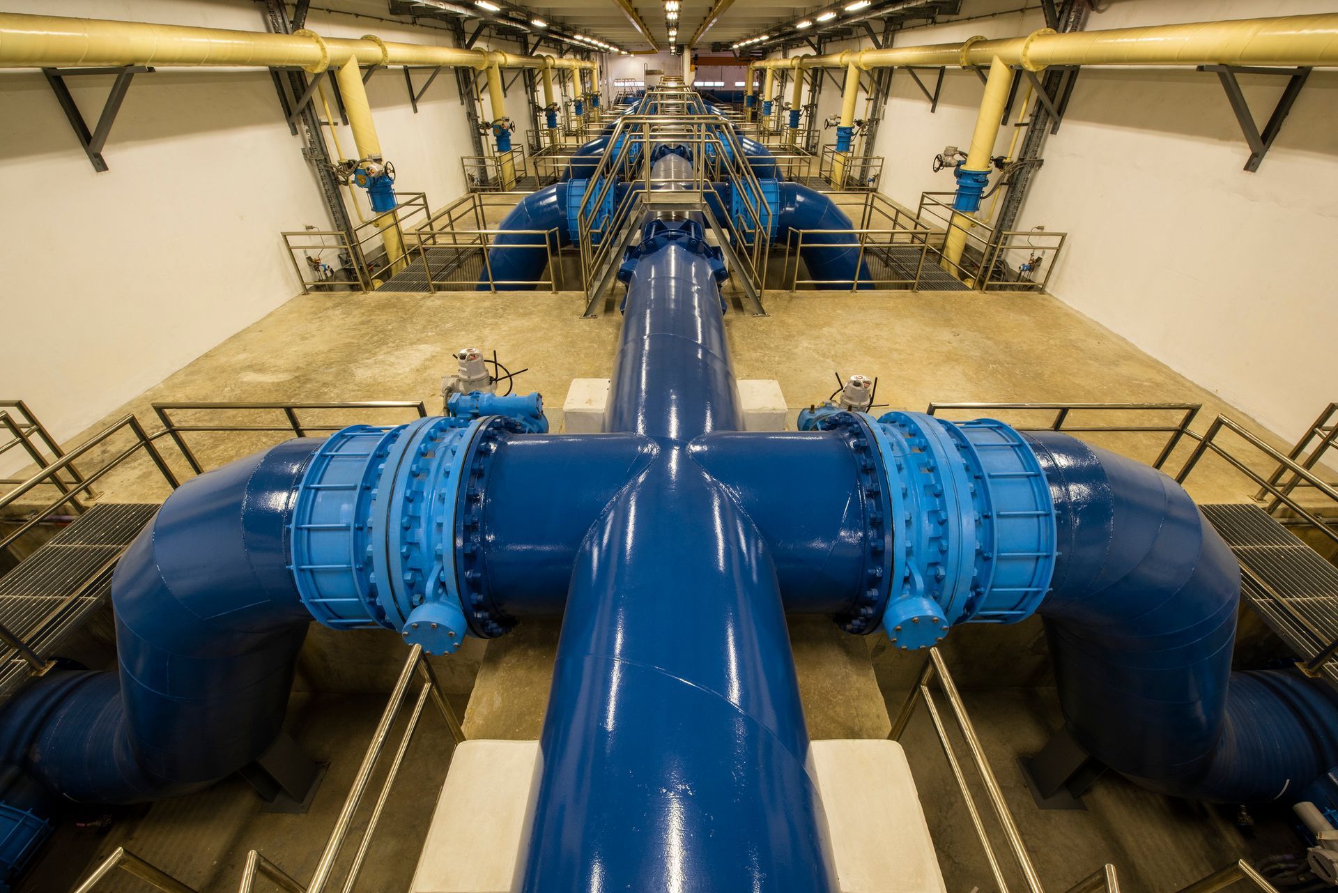 Blue industrial pipes and valves in a utility tunnel with overhead lights and yellow walls