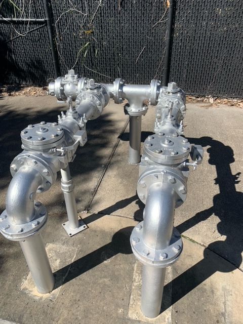 Four silver industrial pipes with valves outdoors on a concrete surface near a black fence