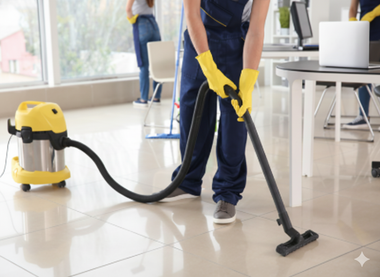Person in yellow gloves vacuuming a tiled office floor with a yellow vacuum cleaner.