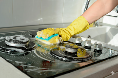 Person wearing yellow glove cleaning a stovetop with a soapy sponge.