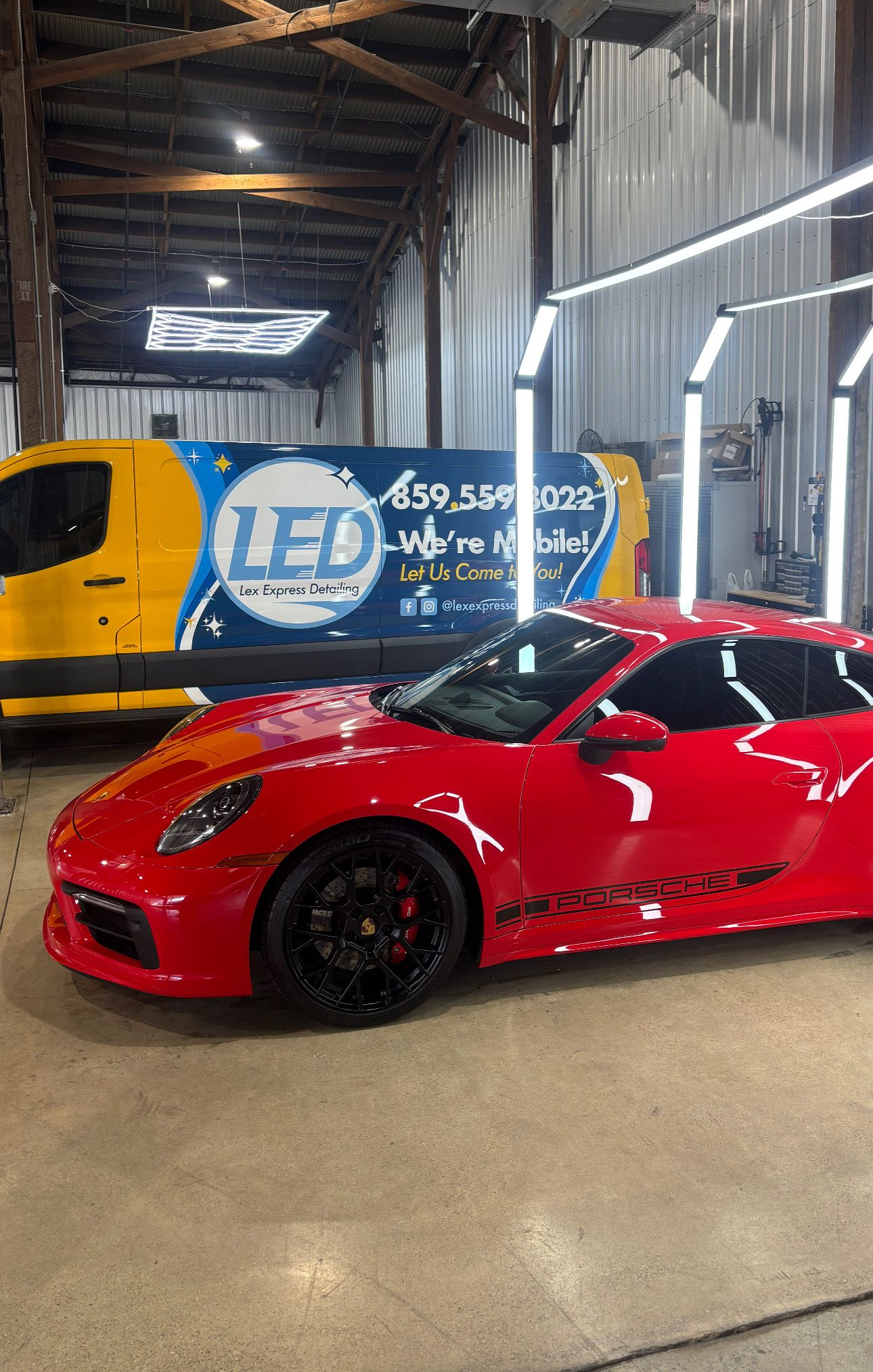 A shiny red Porsche sports car parked inside a garage next to a blue and yellow service van with 