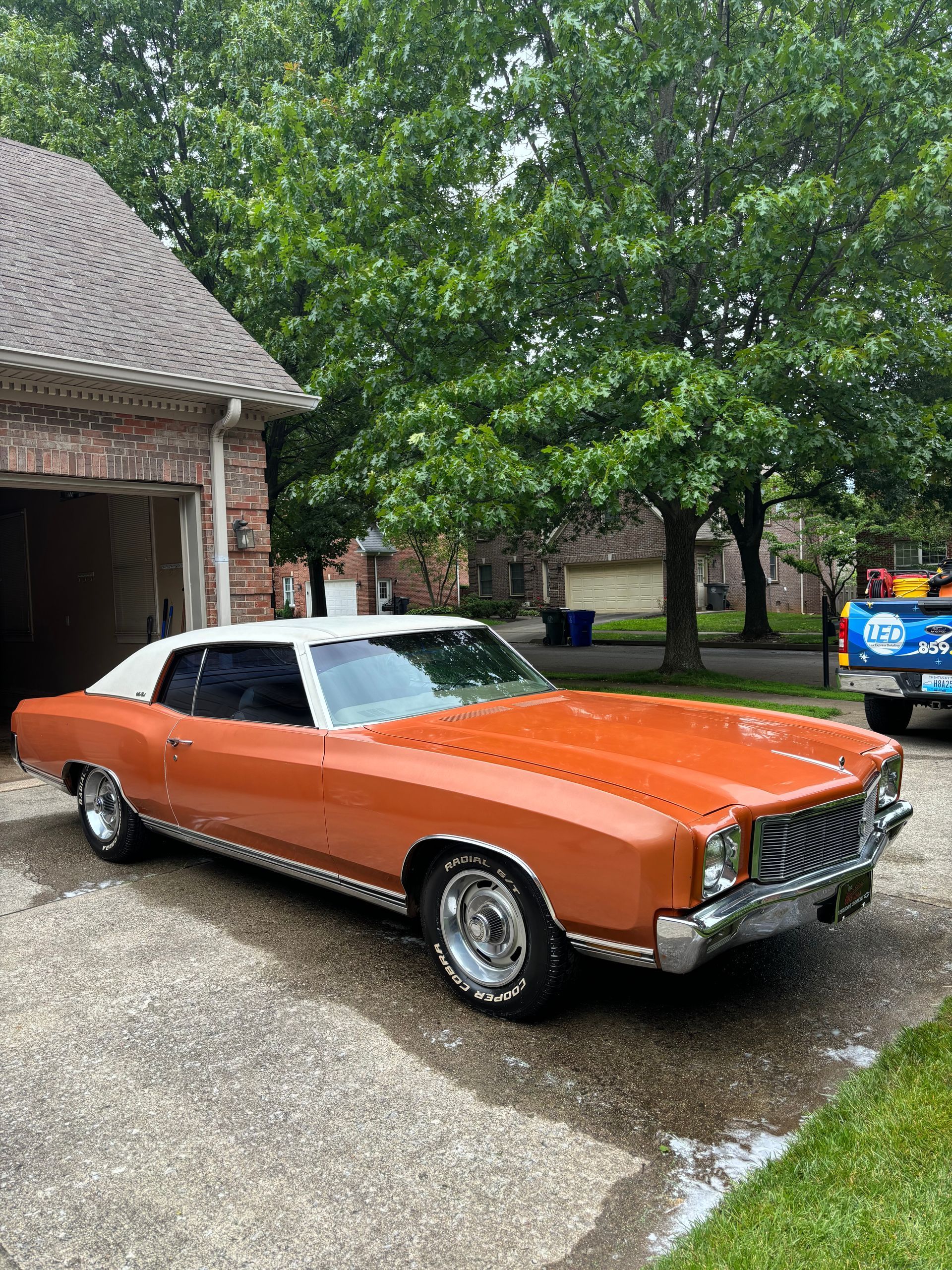 A classic orange and white two-door Chevrolet Monte Carlo parked in a residential driveway on a rainy day.