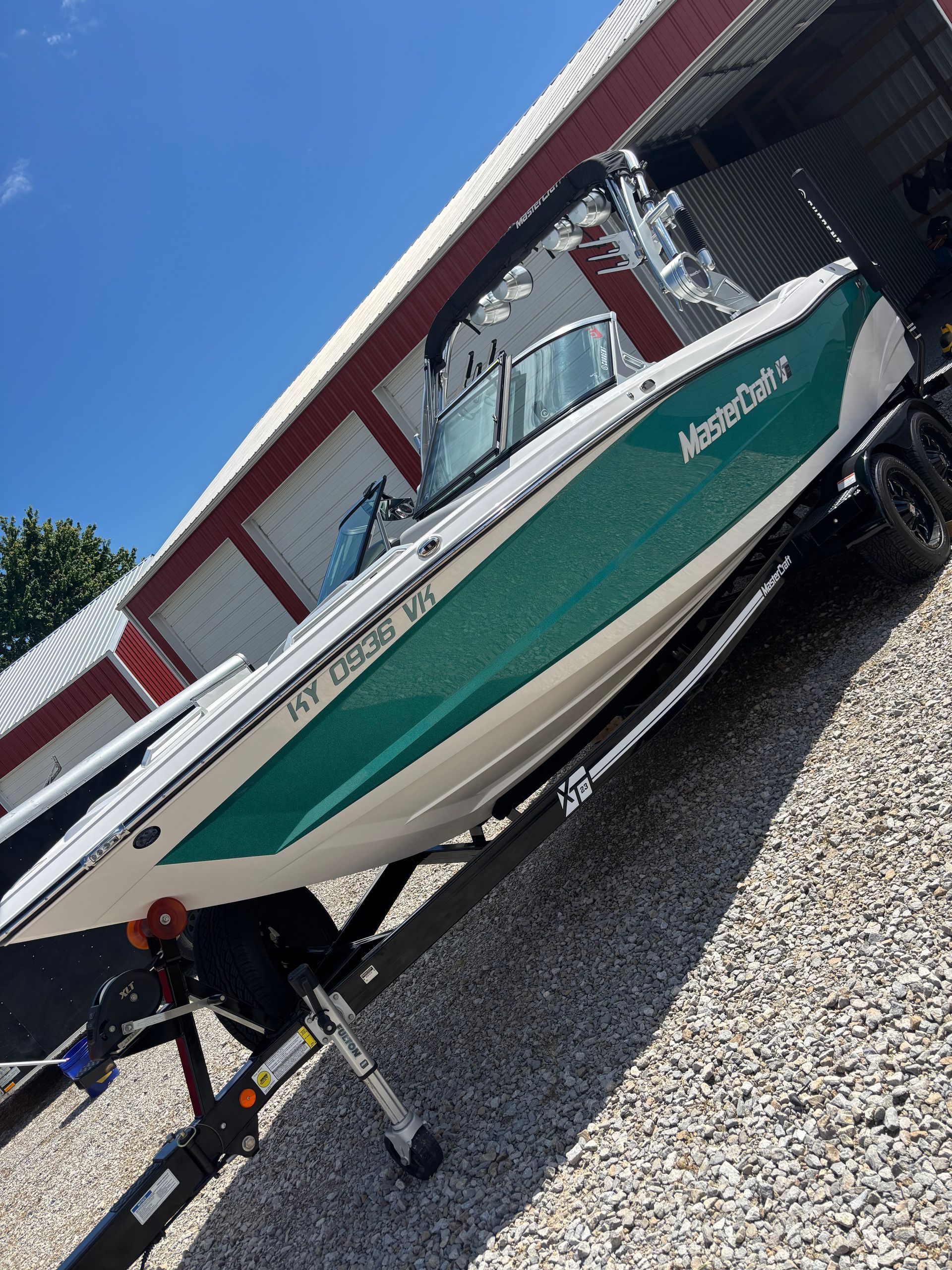 A green and white MasterCraft boat on a trailer parked on a gravel lot next to a building.