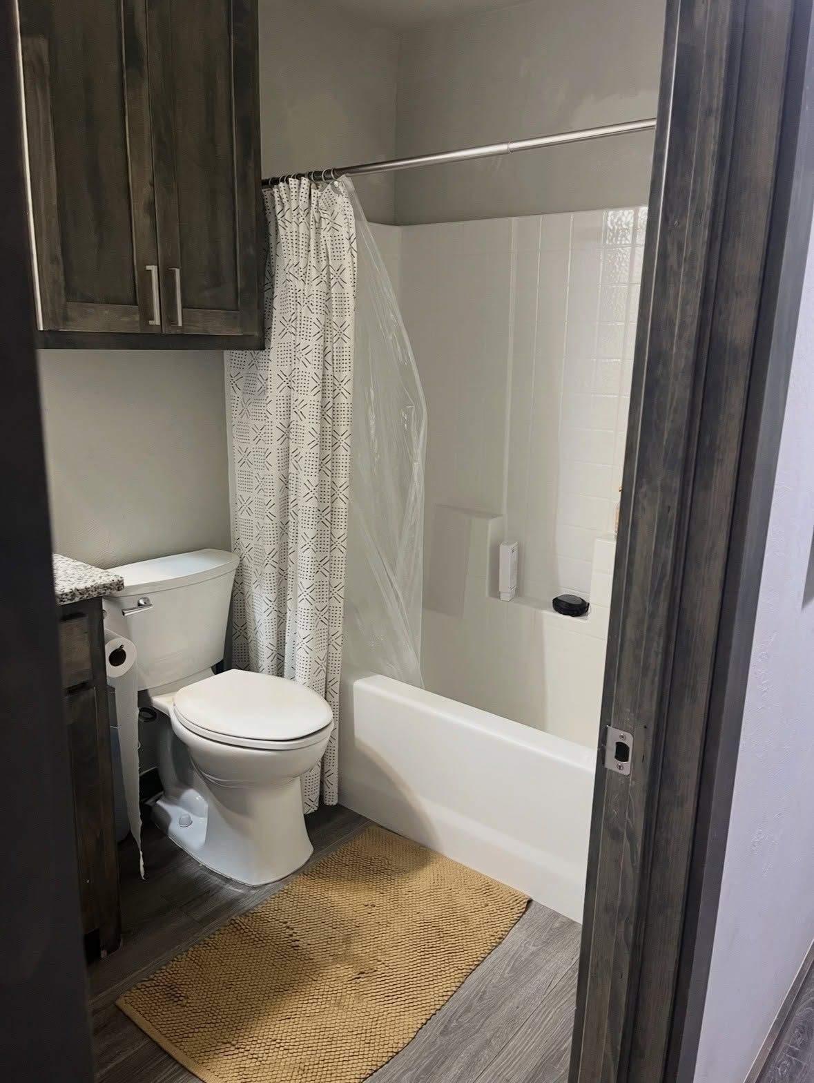 Compact bathroom with toilet, bathtub-shower, dark cabinets, and woven bath mat.