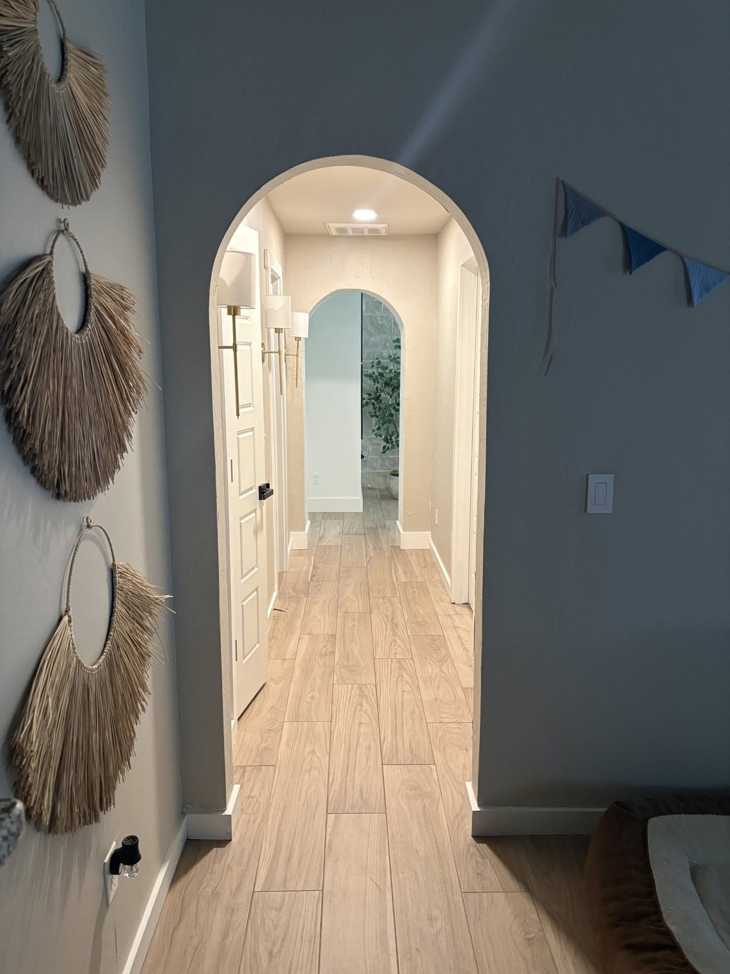 Hallway with arched doorway, light wood floor, and woven wall decor, leading to a bright room.