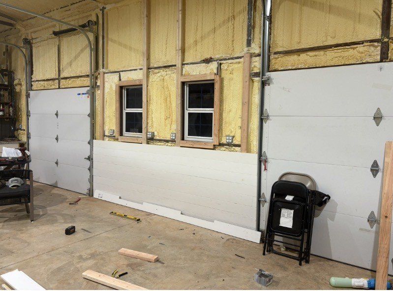 Interior of a garage under renovation with exposed insulation, two windows, and two white overhead doors.