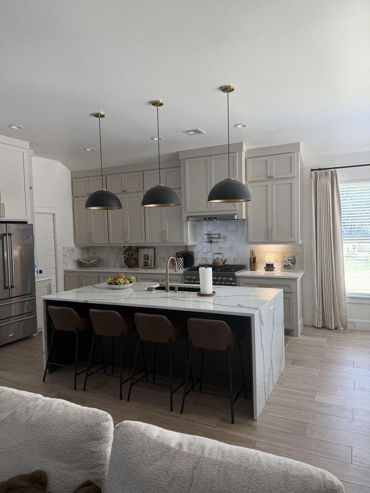 Modern white kitchen with marble island, black pendant lights, and bar stools by a window.