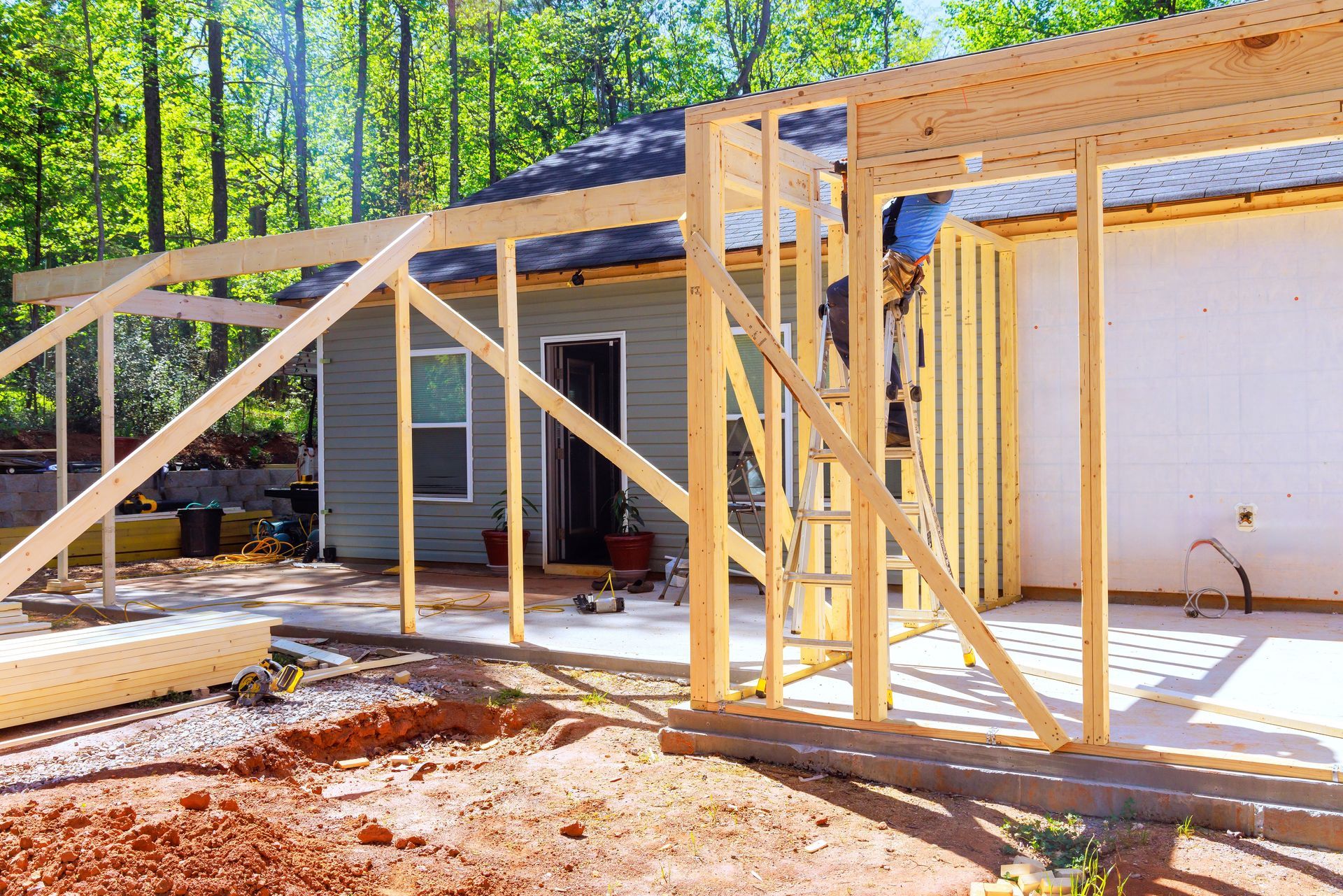 Framed wood structure under construction beside a white garage in a wooded area