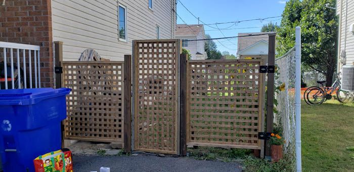 Dans une cour arrière, un portail en treillis de bois est installé entre deux panneaux de clôture identiques, à côté d'une poubelle de recyclage bleue et d'une clôture en grillage.